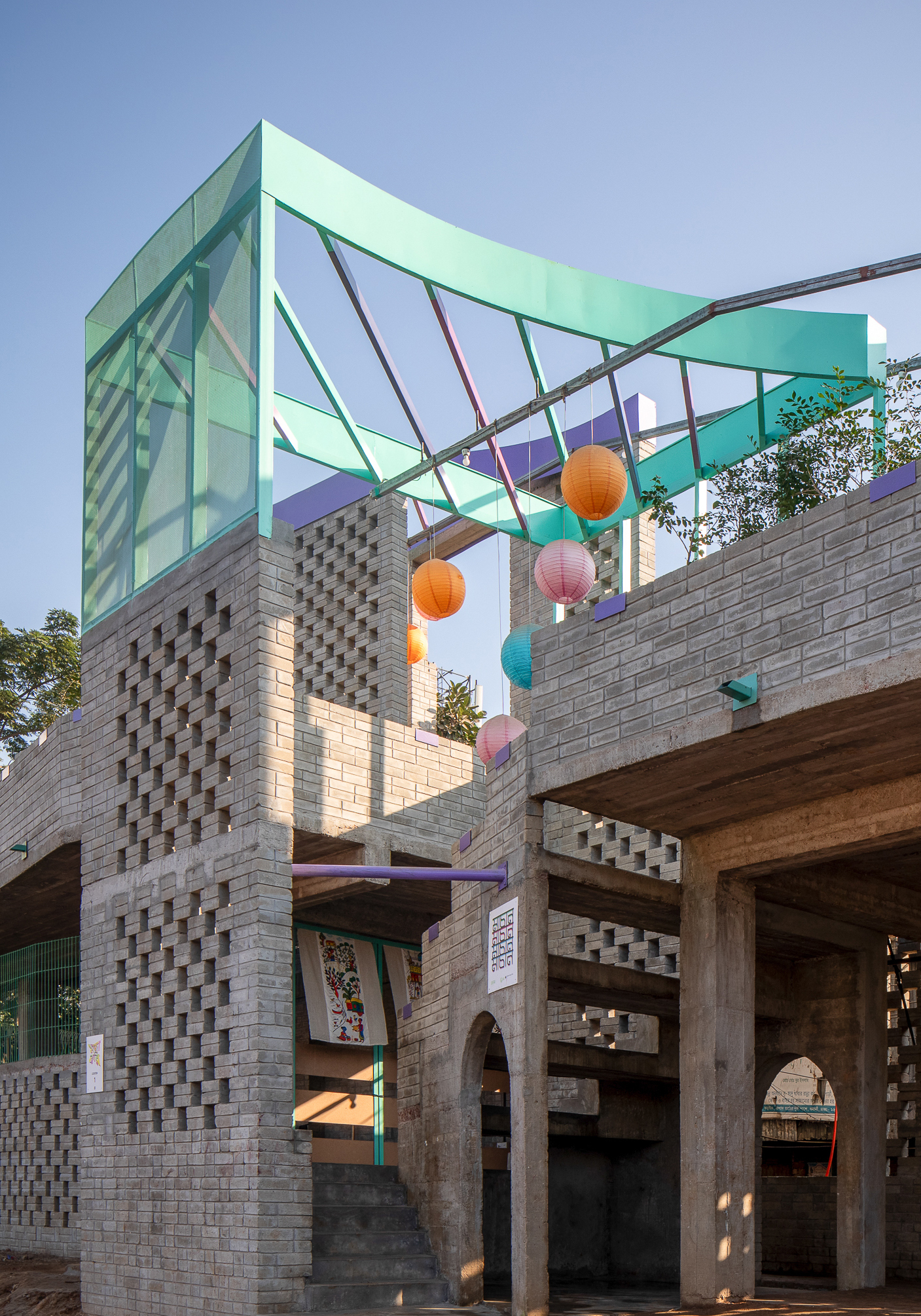 Corner of the perforated brick tower topped with a curved teal steel canopy and hanging paper lanterns under a clear blue sky