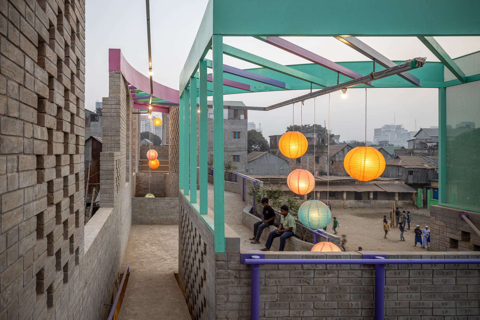 Rooftop terrace with teal frames, purple railings and glowing paper lanterns as two seated figures look over the low neighborhood at dusk