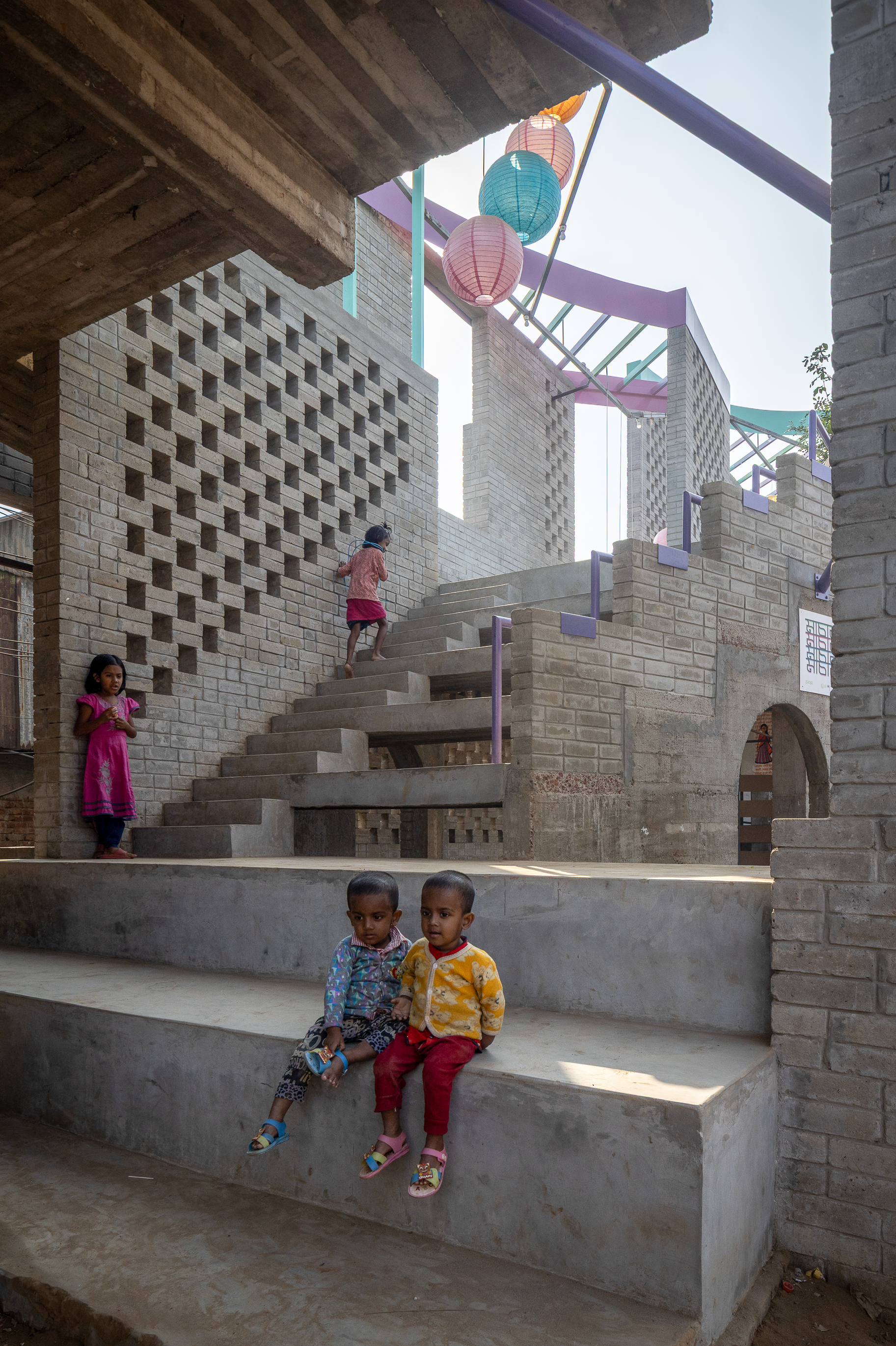 Two small children sit on concrete steps in front of a brick lattice wall and stair, with a girl climbing above in sunlight