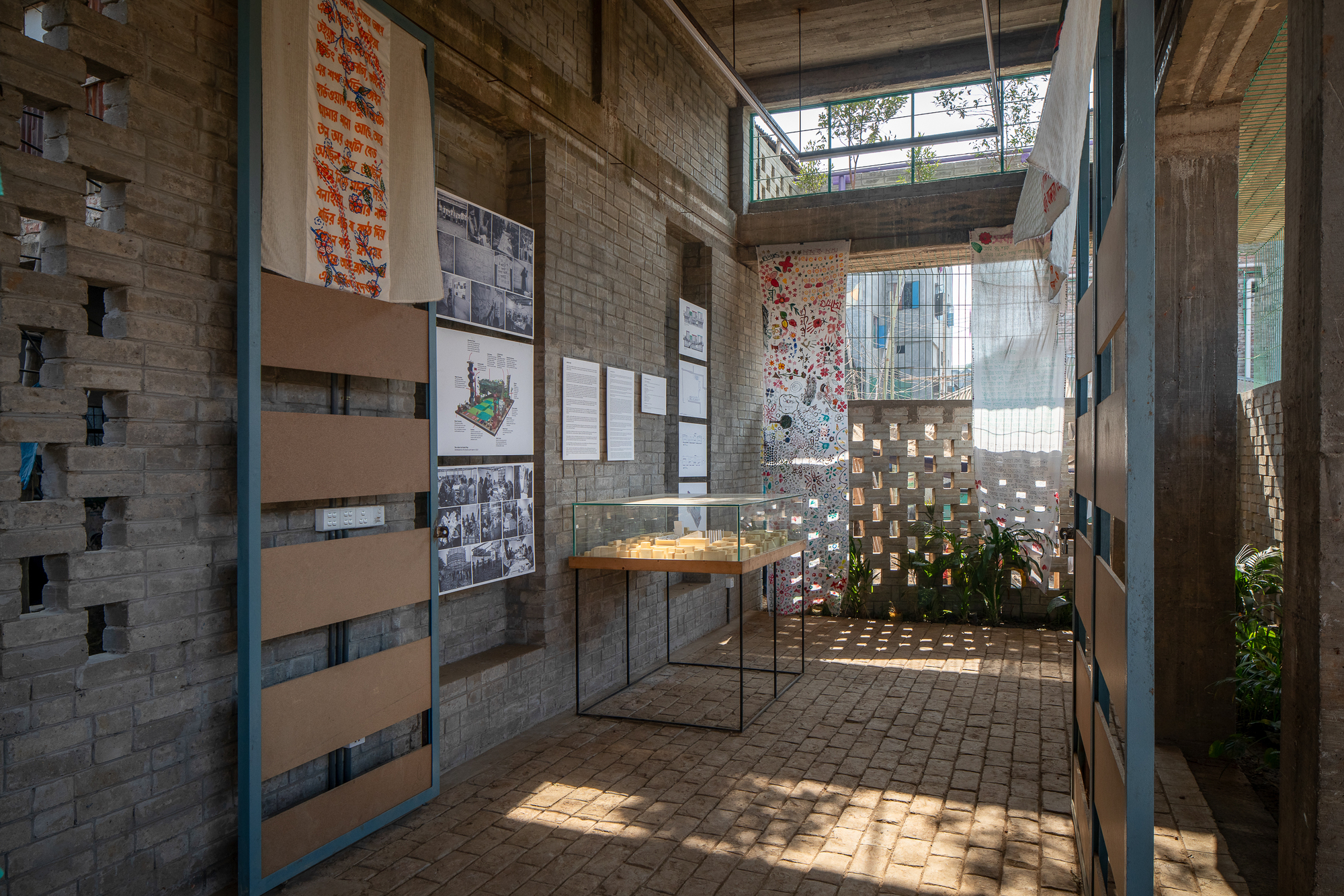 Interior gallery corridor with brick floor, display vitrine, pinned photographs and hanging textiles lit by sunlight filtering through perforated brick walls