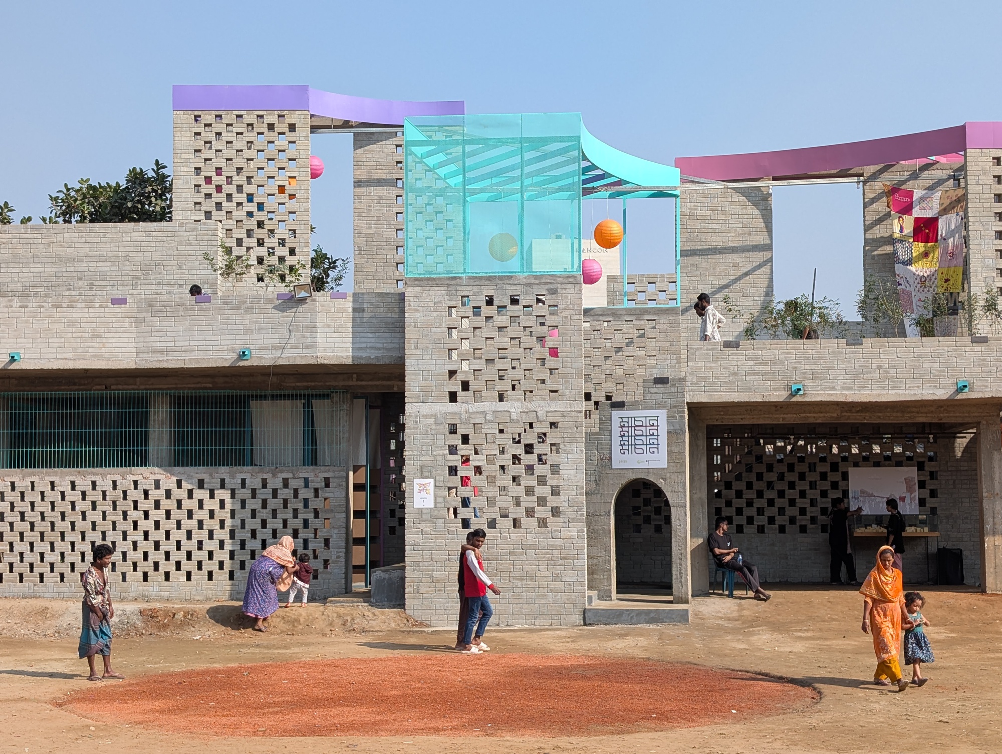 Perforated brick facade with a glass rooftop pavilion and colored canopies, women and children crossing the red earth courtyard in clear daylight