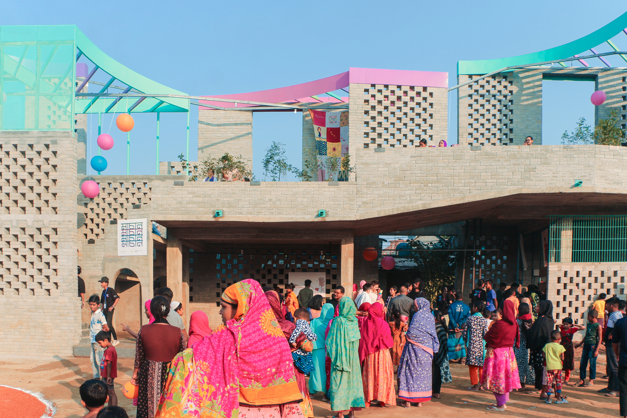 Crowd of women in bright saris gathered beneath a brick porch with green and pink curved roof canopies and hanging paper lanterns