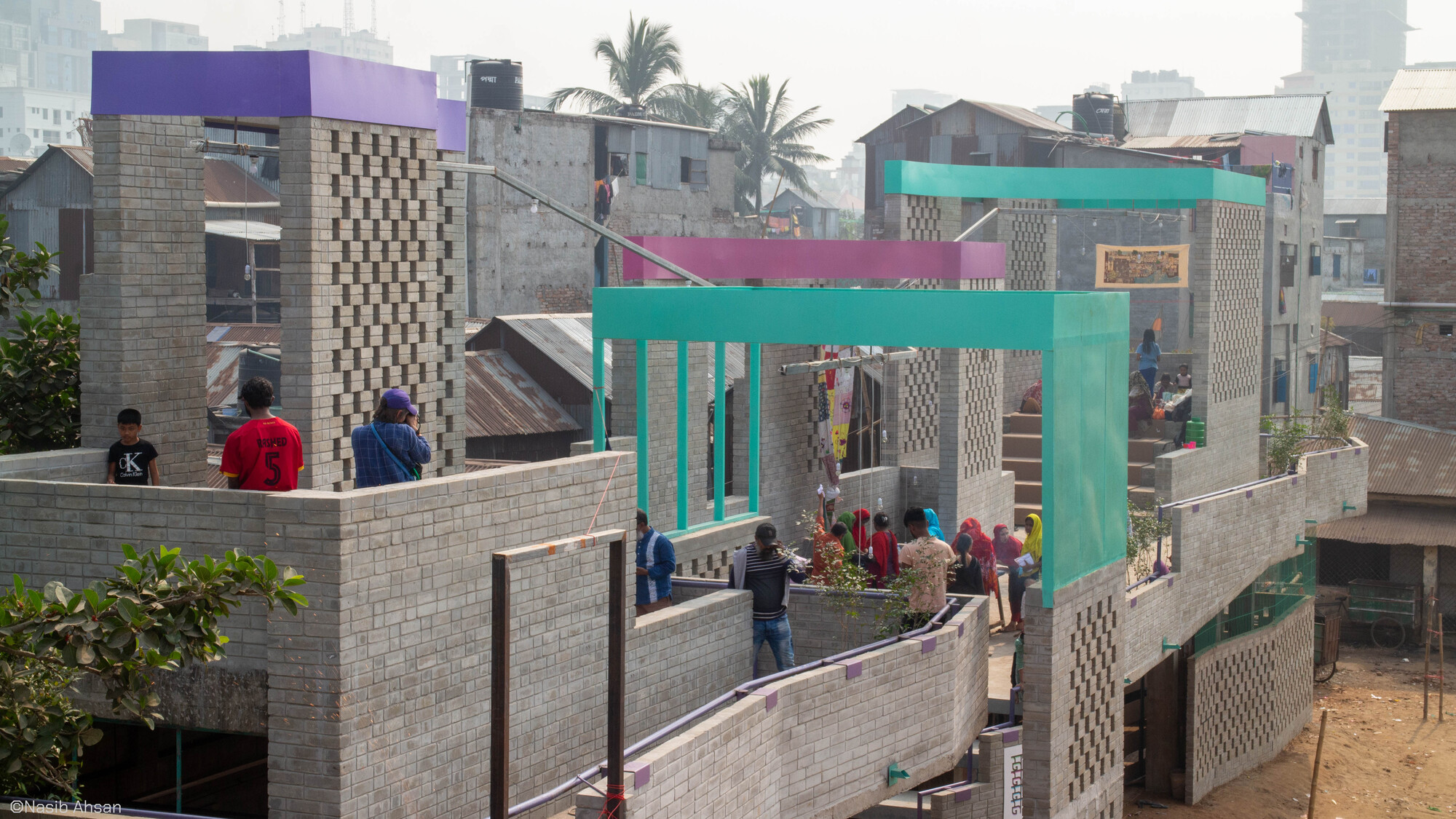 Elevated view of brick terraces with perforated screen walls and turquoise and purple frames, visitors standing along the parapet in hazy light