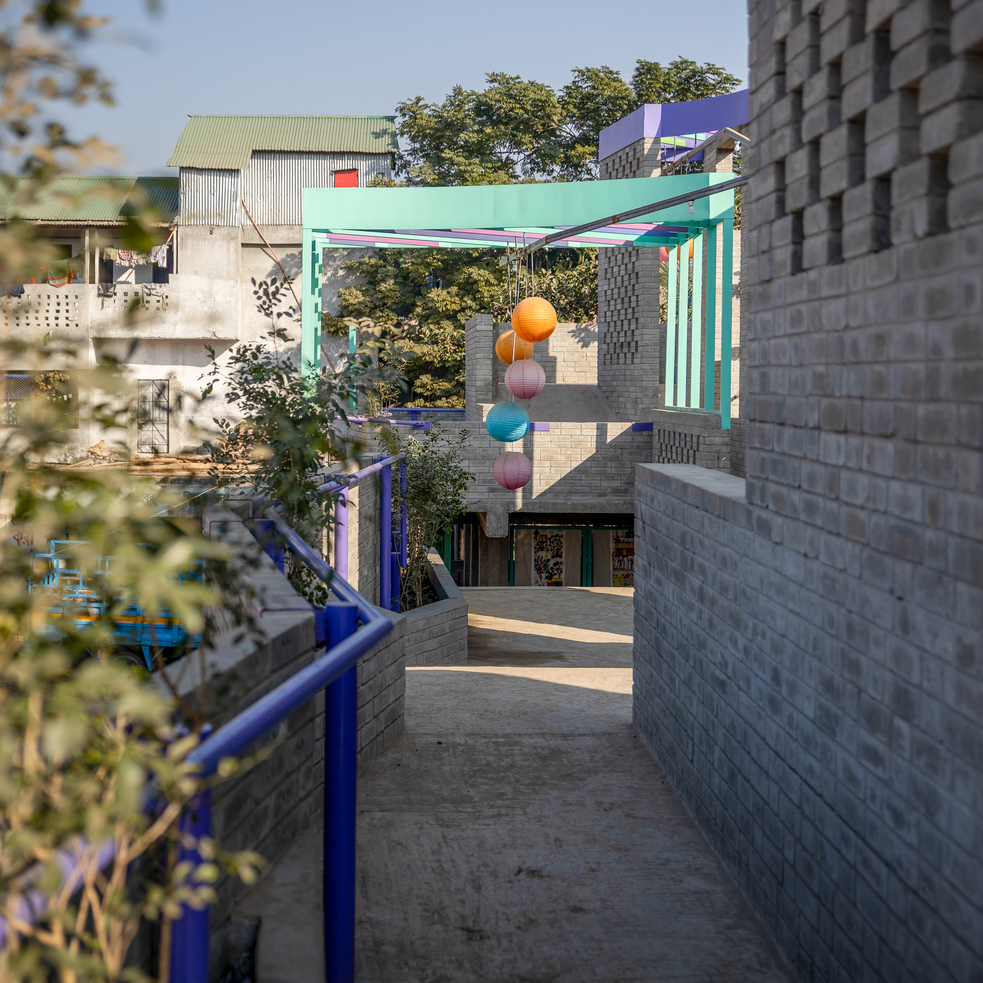 Narrow concrete walkway between brick walls with blue railings, leading to a teal pergola and hanging paper lanterns in low sun