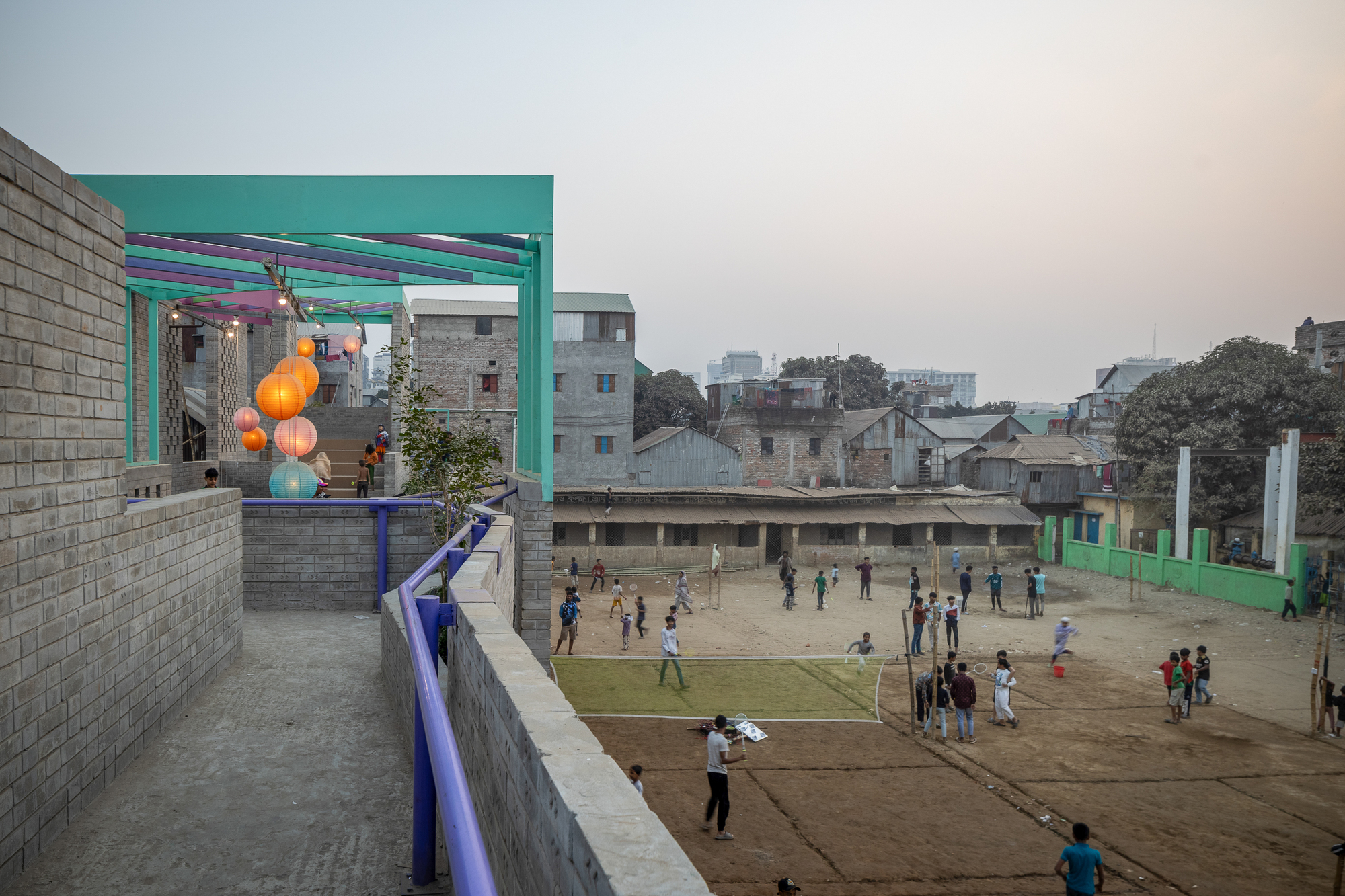 Brick terrace with blue railing overlooking a dirt field where children play volleyball and games at dusk, lanterns glowing under a teal canopy