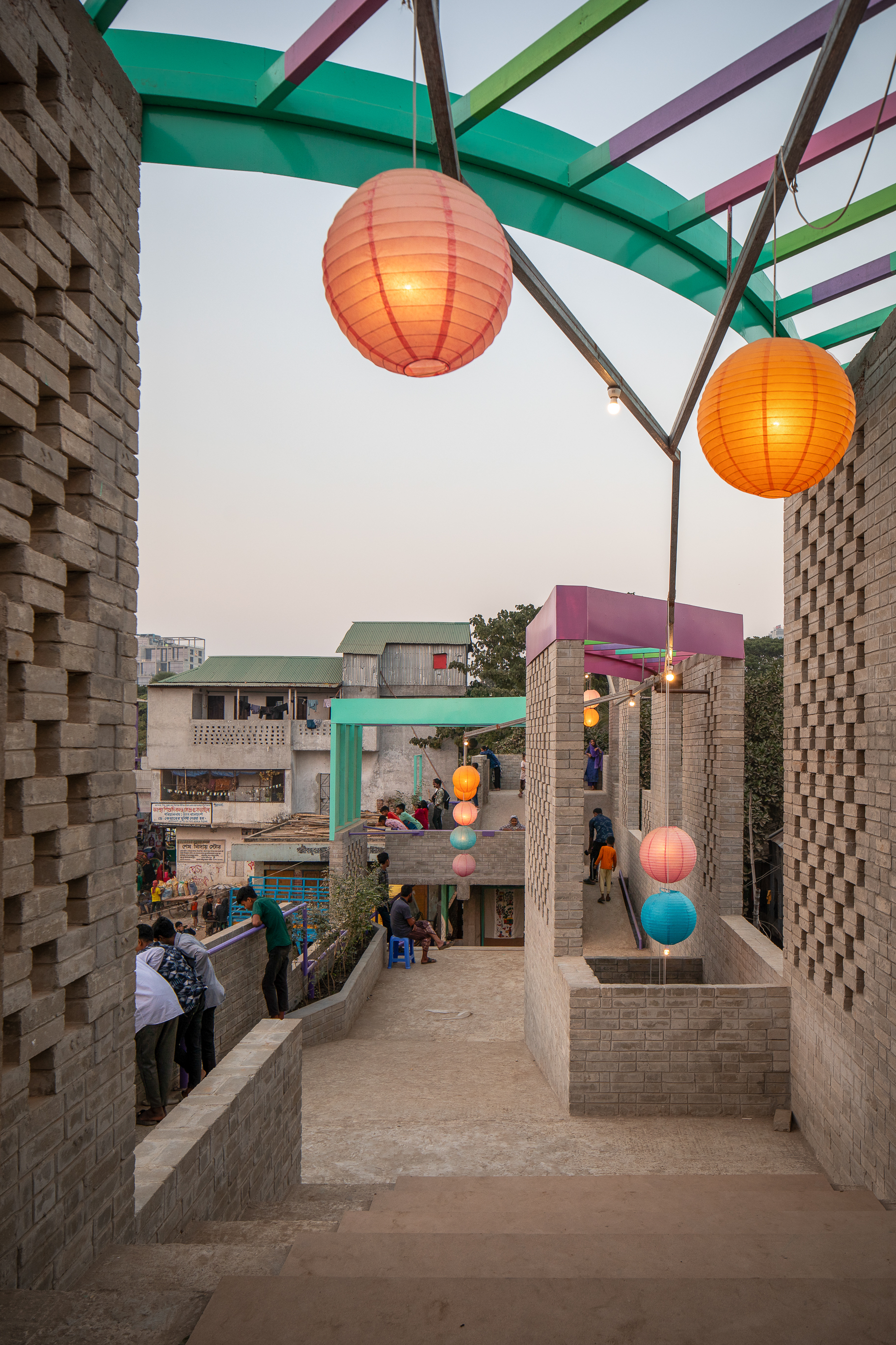 Brick stairway climbing toward glowing orange and blue paper lanterns under a curved green canopy, with a few people gathered at dusk