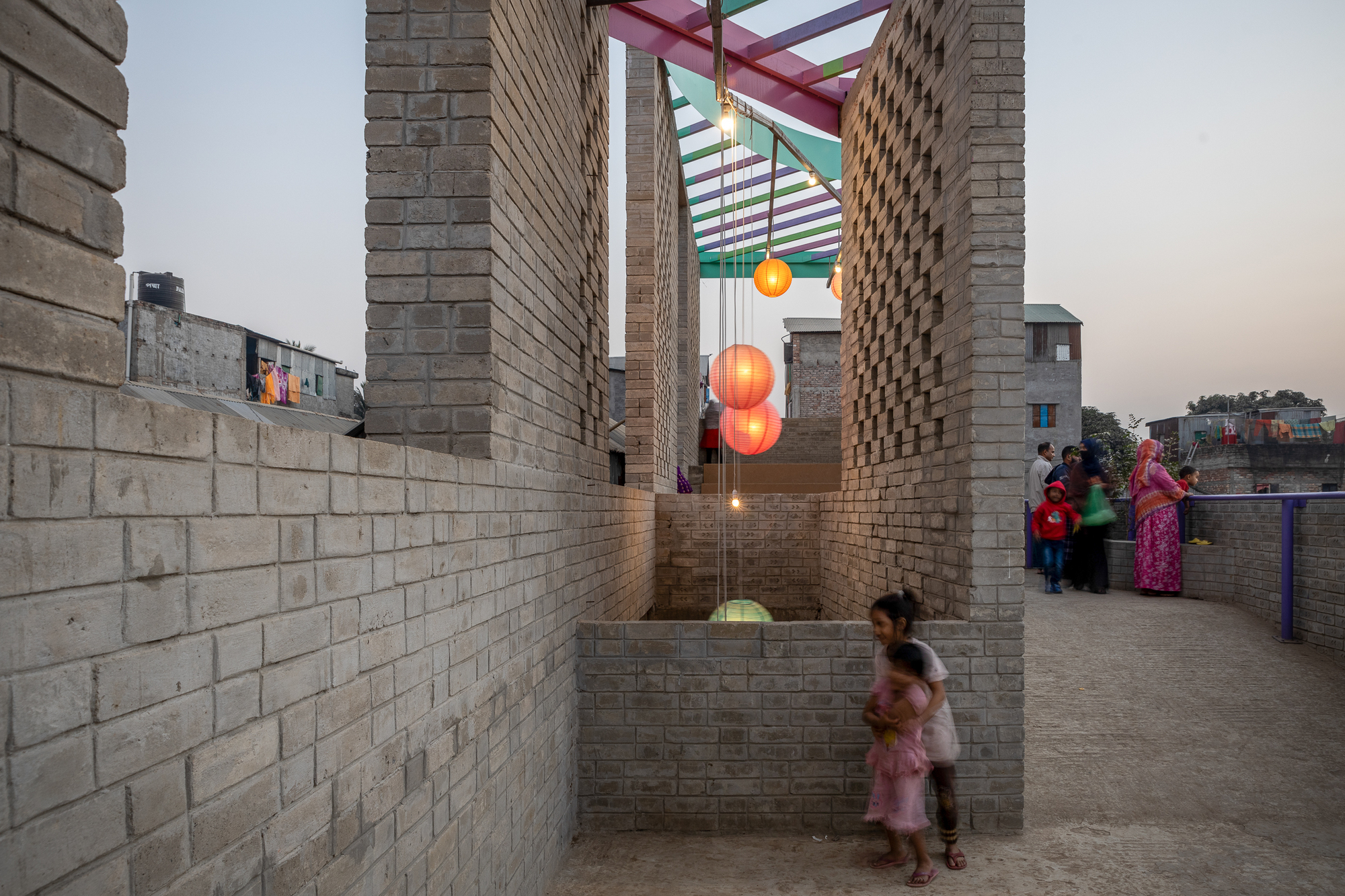 Narrow brick passage between perforated walls with glowing lanterns under a colorful pergola, a child running past at dusk