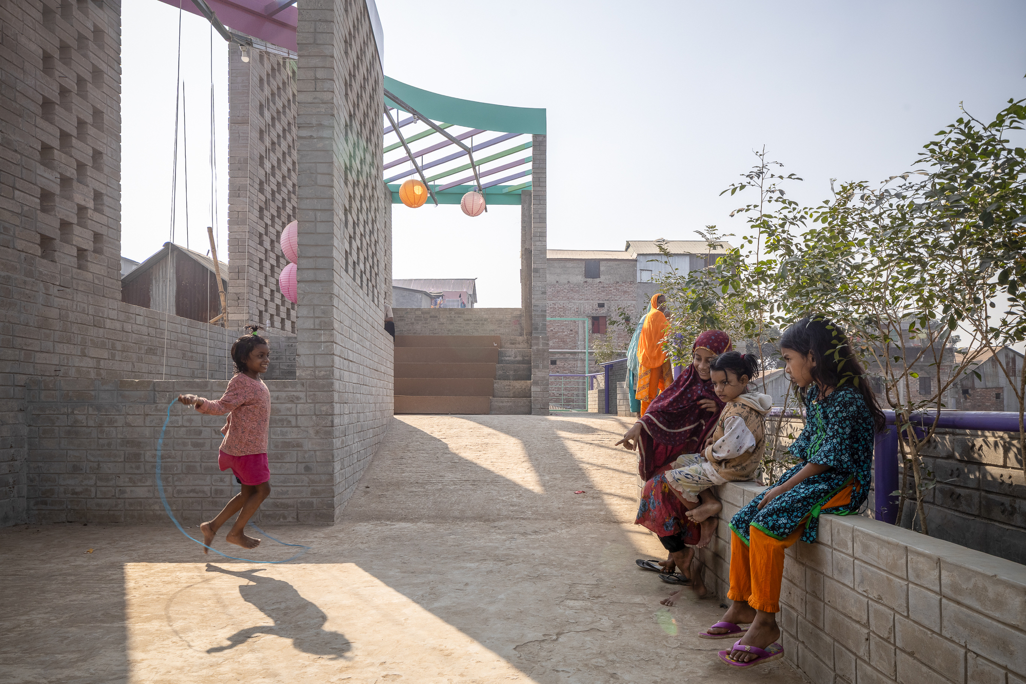 Girl skipping rope on a sunlit brick terrace while a woman and children sit on a low wall beside a leafy shrub