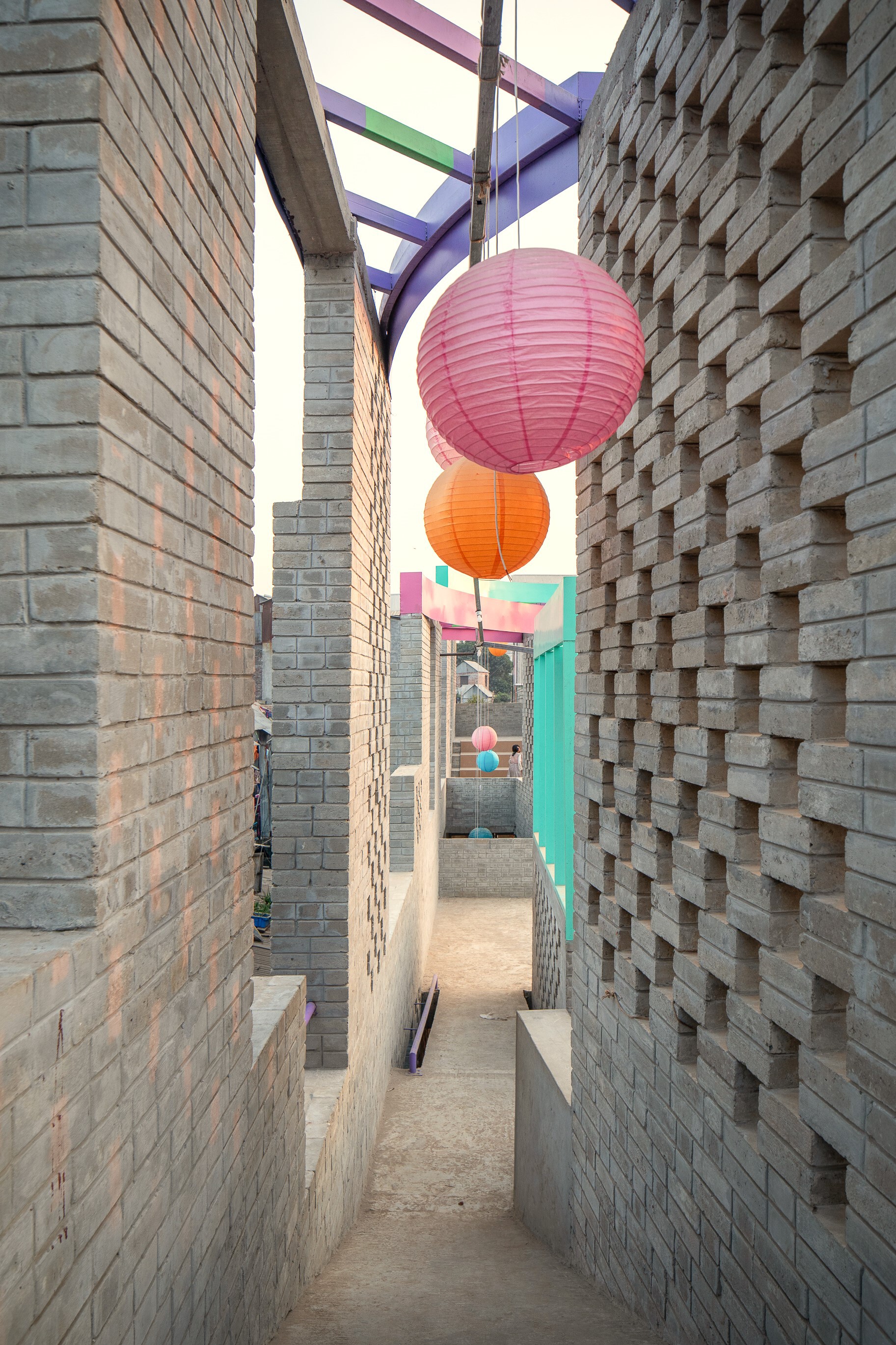 Slim alley between perforated brick walls with pink and orange paper lanterns hanging beneath a purple pergola frame