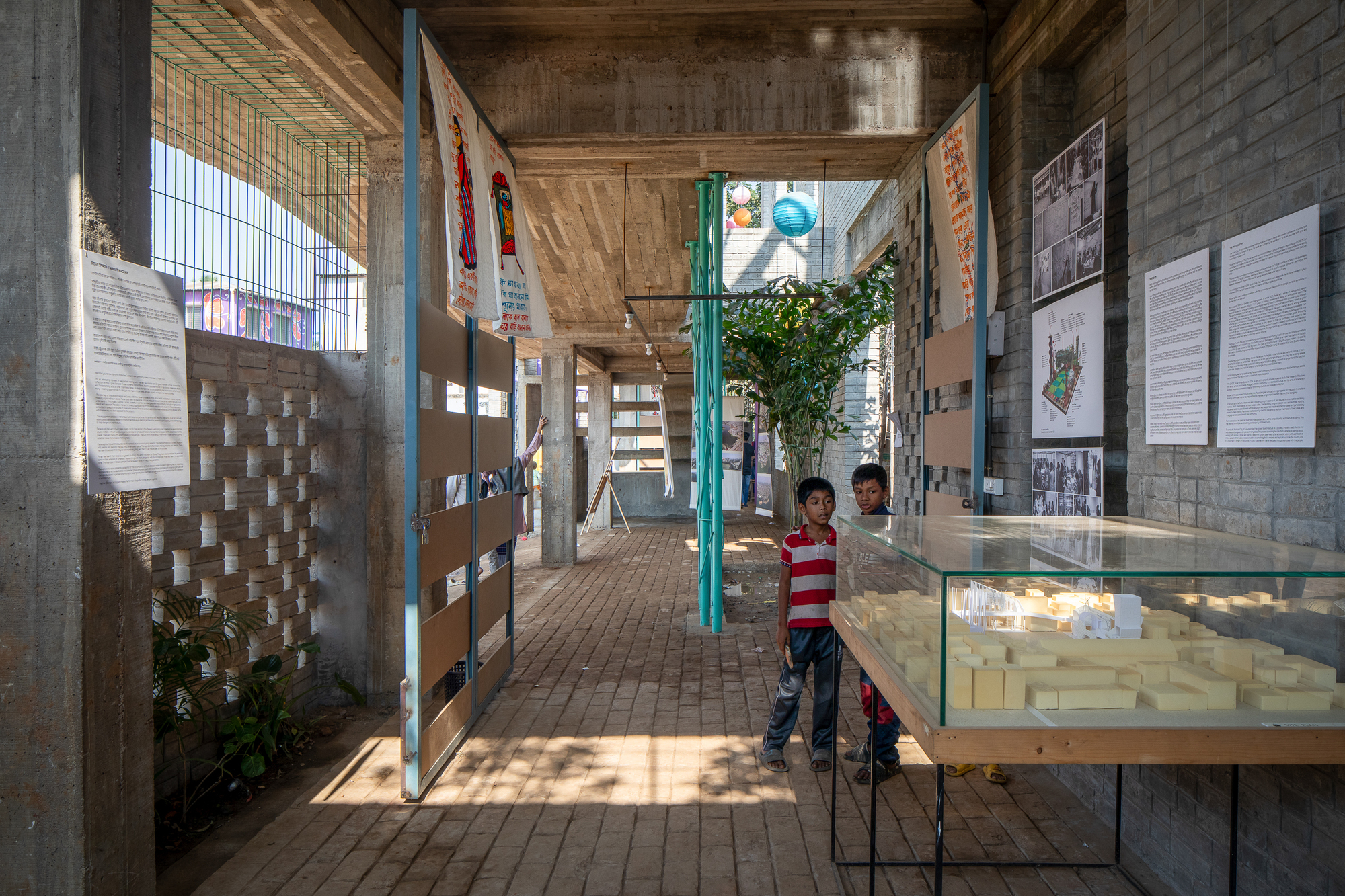 Covered brick walkway with concrete ceiling, pinned exhibition panels and a glass-topped model case, two boys standing in dappled daylight