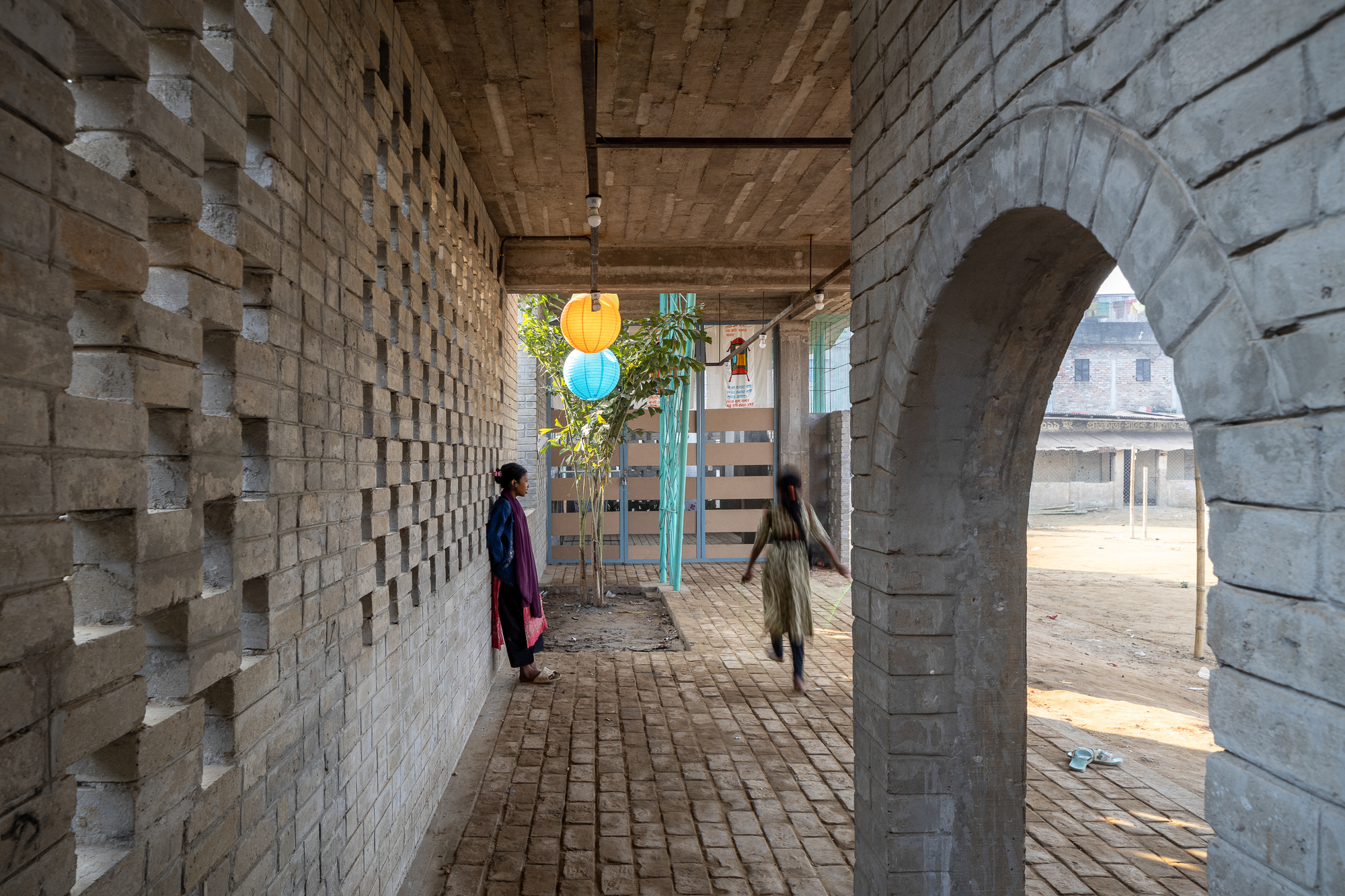 Covered brick passage with a perforated brick wall and rounded arch, a child running past a hanging orange lantern in warm daylight