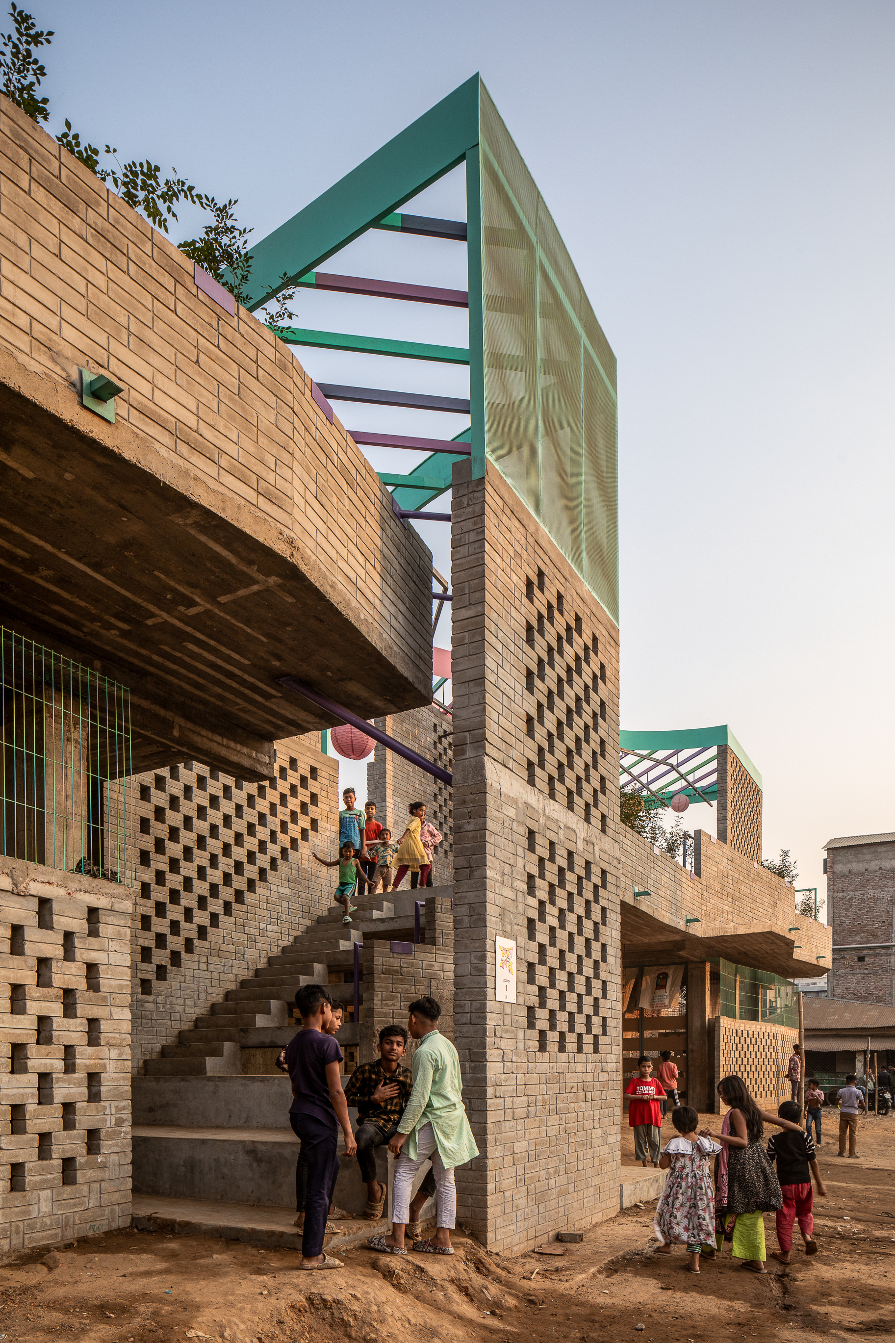 Brick facade with perforated screen walls, an open stair and a green steel frame above, children gathering at dusk