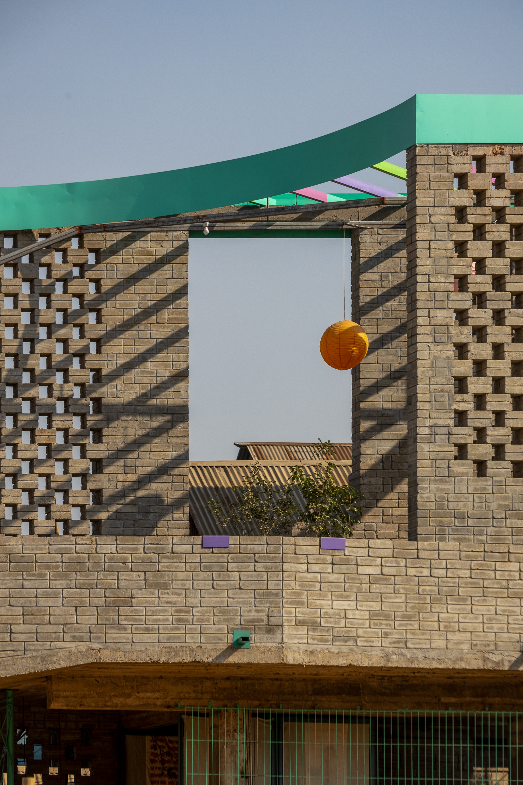 Perforated brick walls frame a square opening with a hanging orange lantern under a curved green roof edge in strong sun