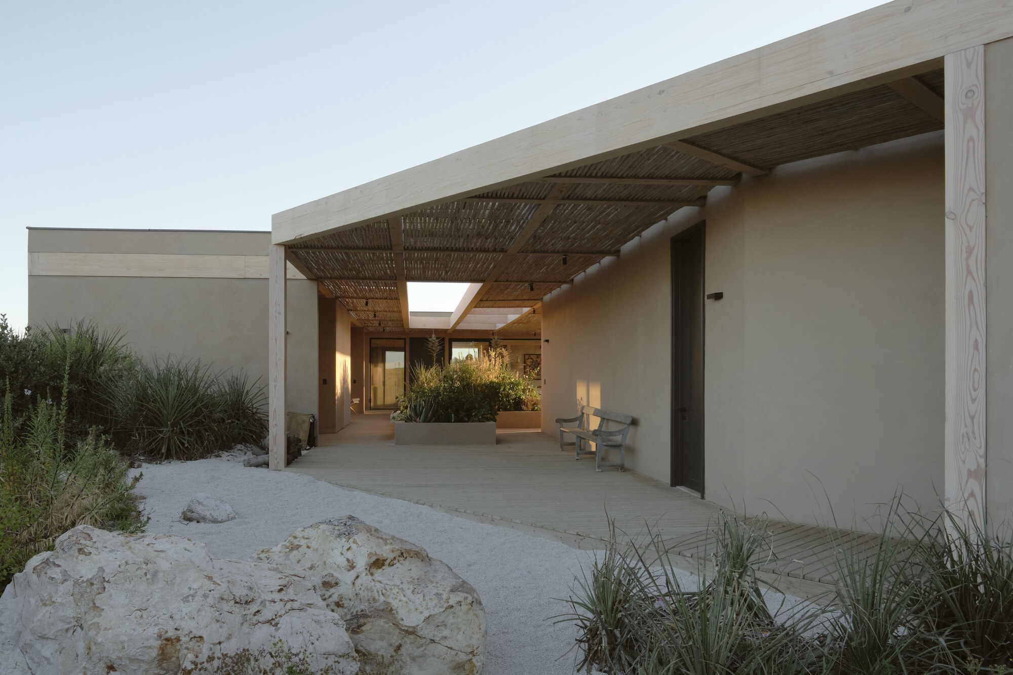 Covered walkway with pale timber posts and a woven cane ceiling, a bench against the earthen wall and white gravel in warm evening light