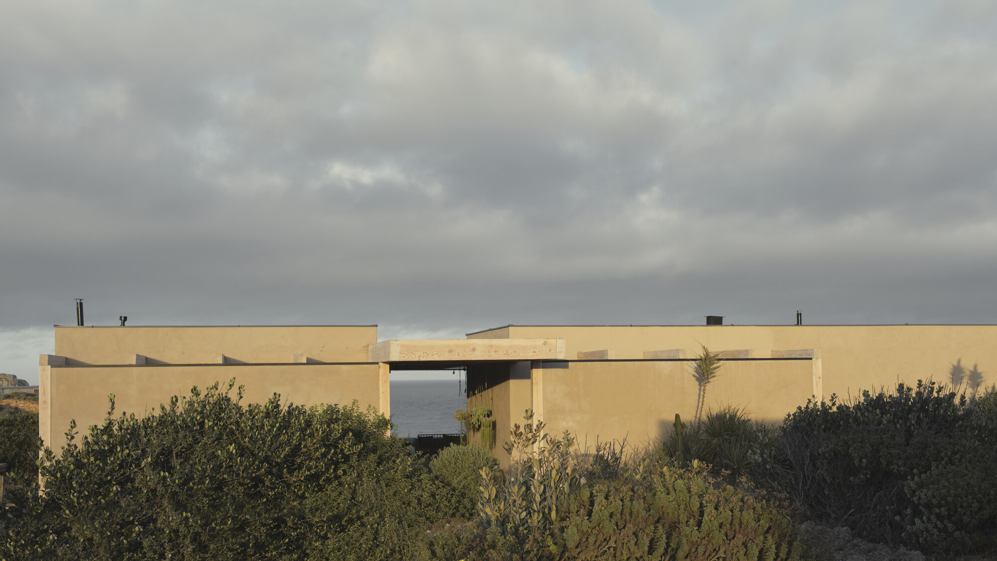 Low earth-toned volumes with an open gap framing the sea, seen over coastal scrub under heavy grey clouds