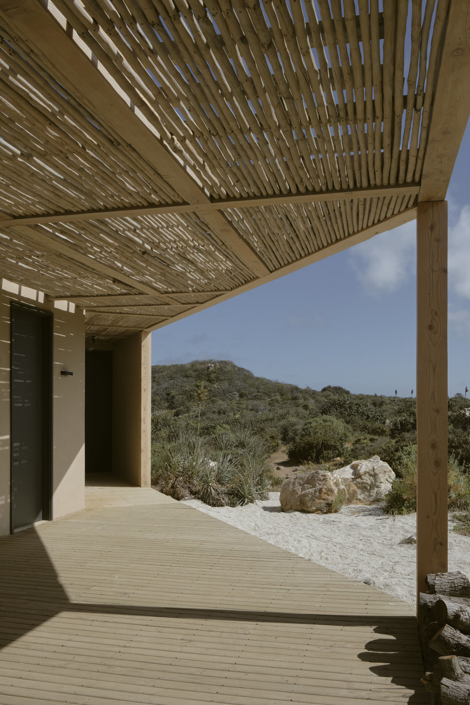Timber deck beneath a lattice of split cane overhead, with boulders and white sand beyond in bright midday sun