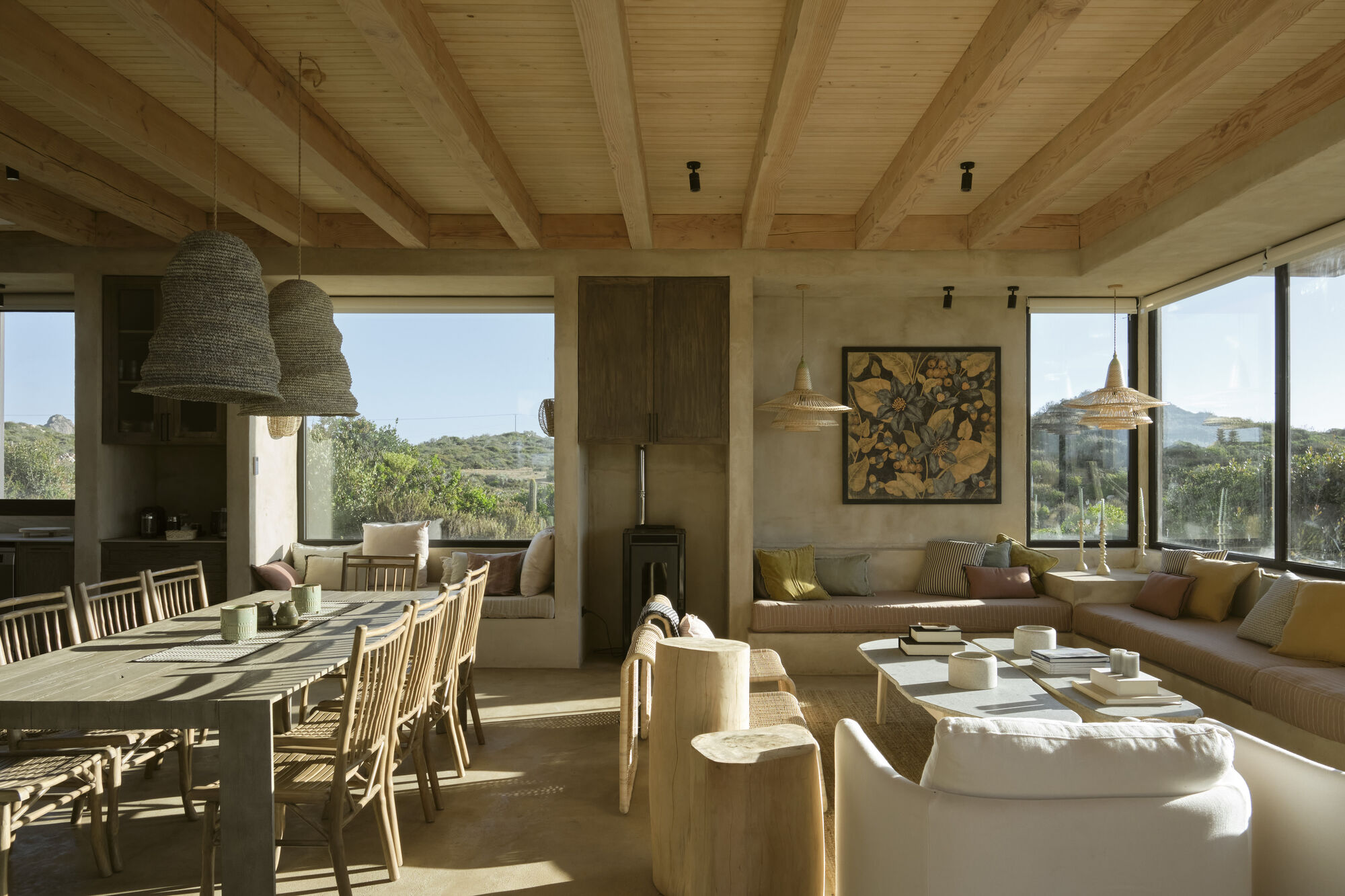 Open living and dining room with exposed timber beams, wicker pendant lamps and a built-in sofa facing hillside windows