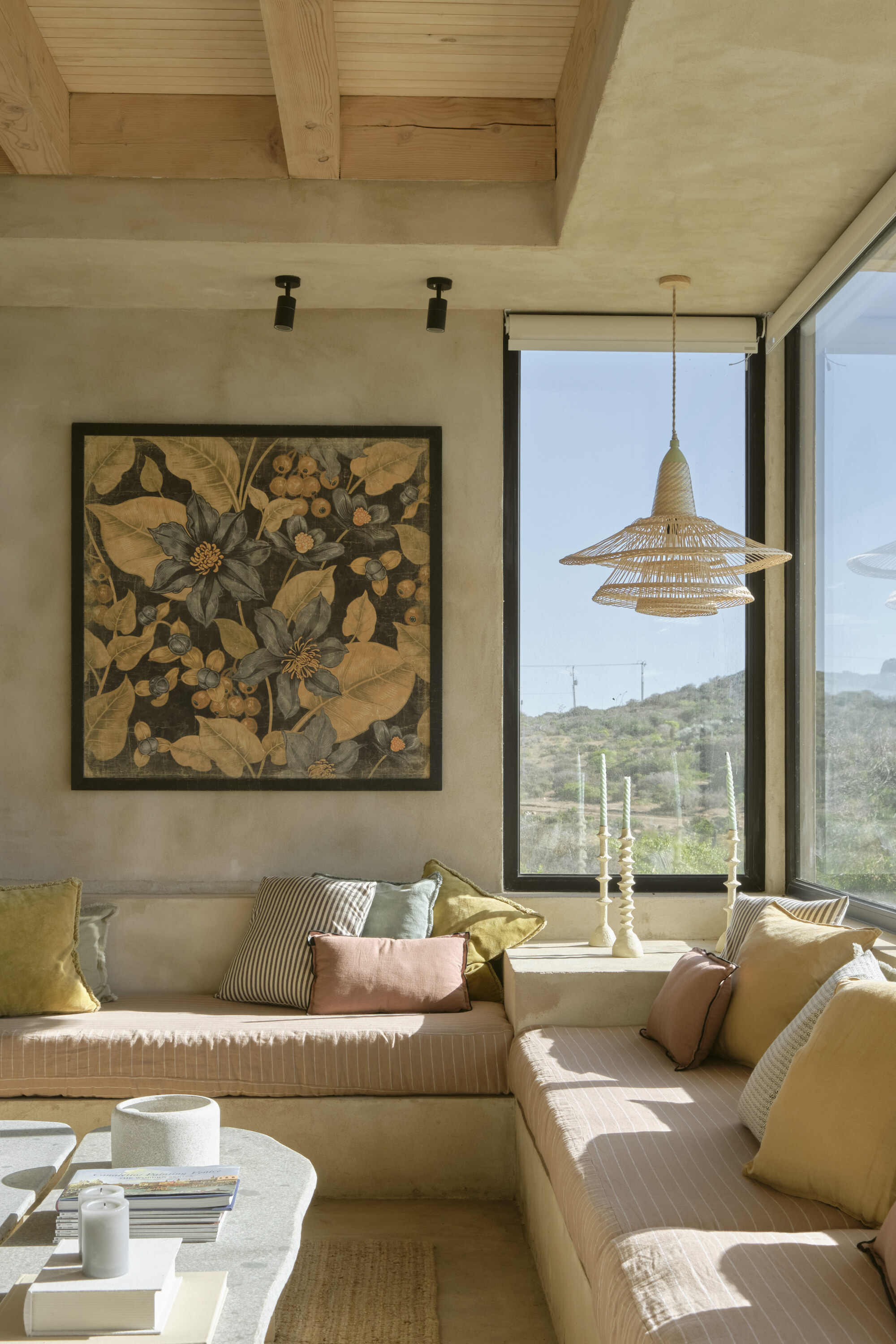 Built-in cushioned bench beneath a large botanical panel, with a woven pendant lamp and a window onto sunlit hills