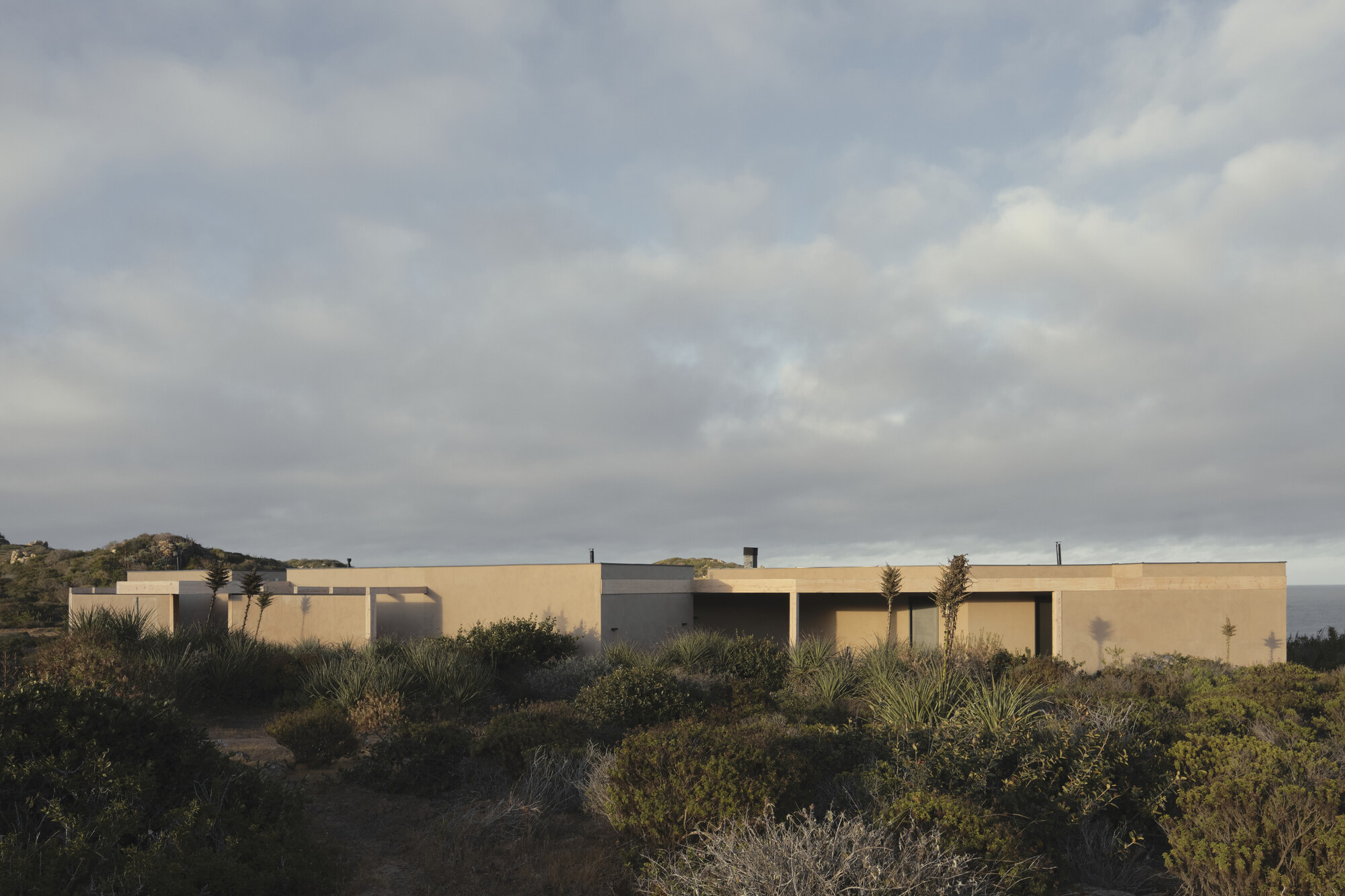 Long single-storey house of plastered walls and flat roofs behind native shrubs, lit by low sun under layered clouds