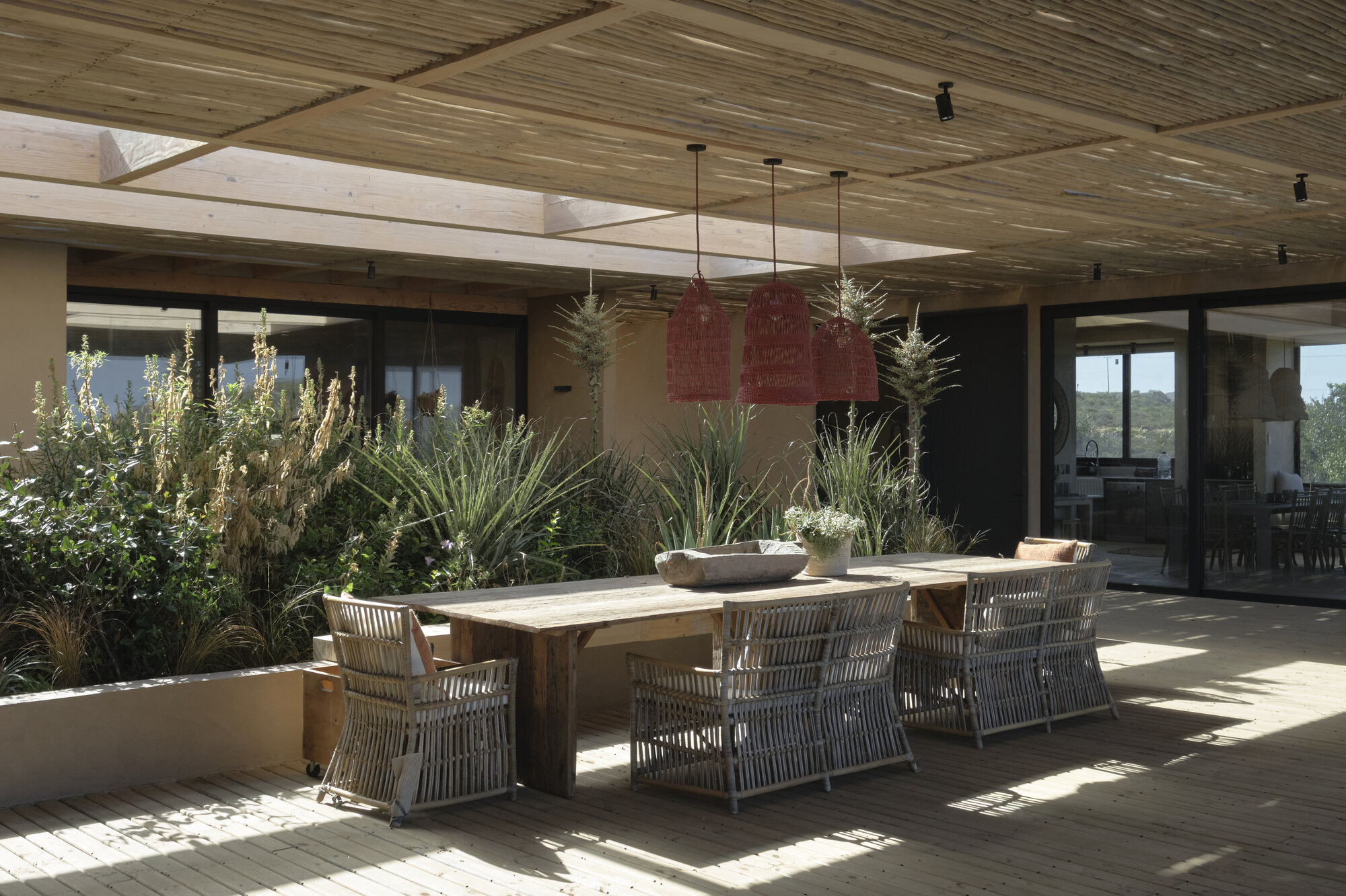 Covered deck with a long timber table, wicker chairs and red pendant lamps beneath a reed-slatted roof, dappled with sunlight