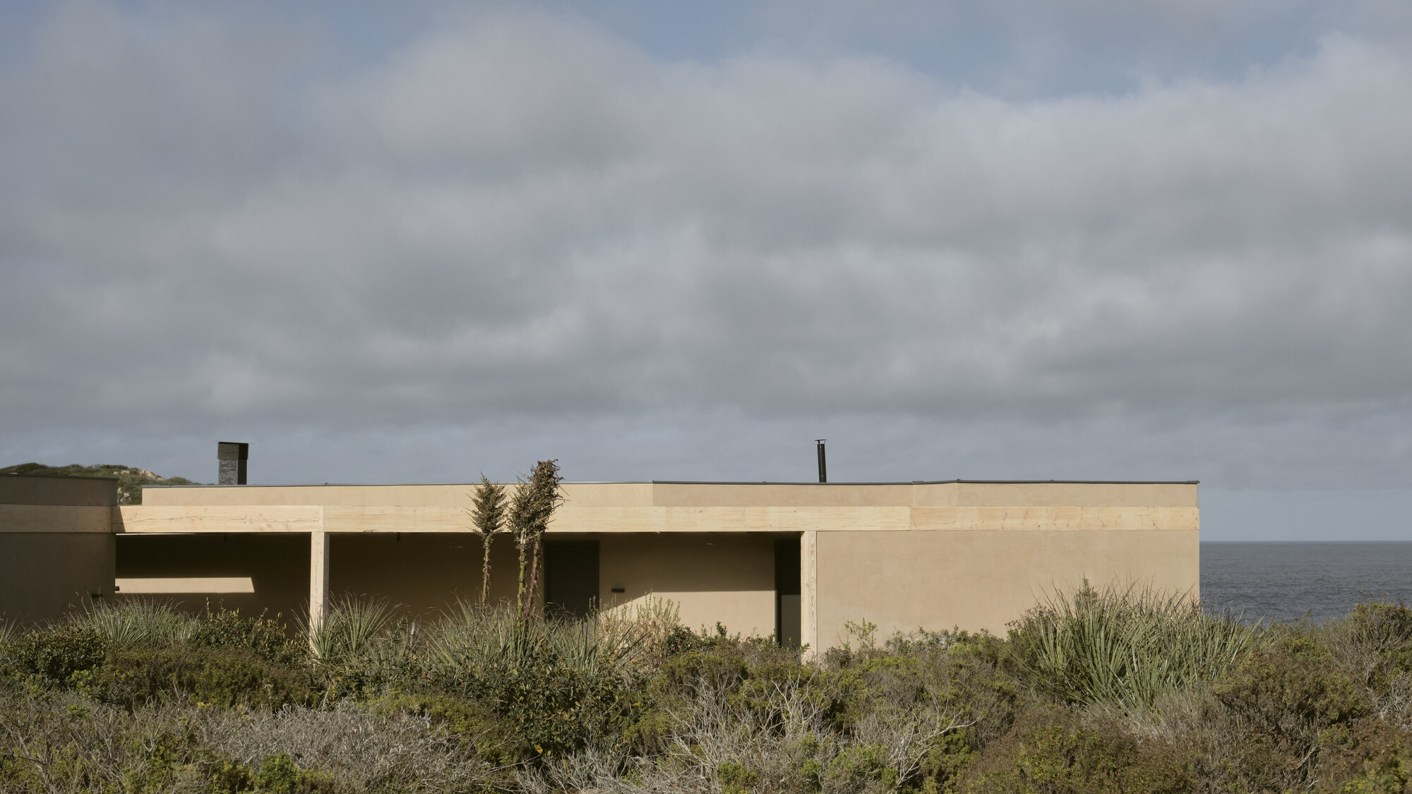 Low flat-roofed house of sand-colored walls seen over coastal scrub, with the sea behind under heavy grey clouds