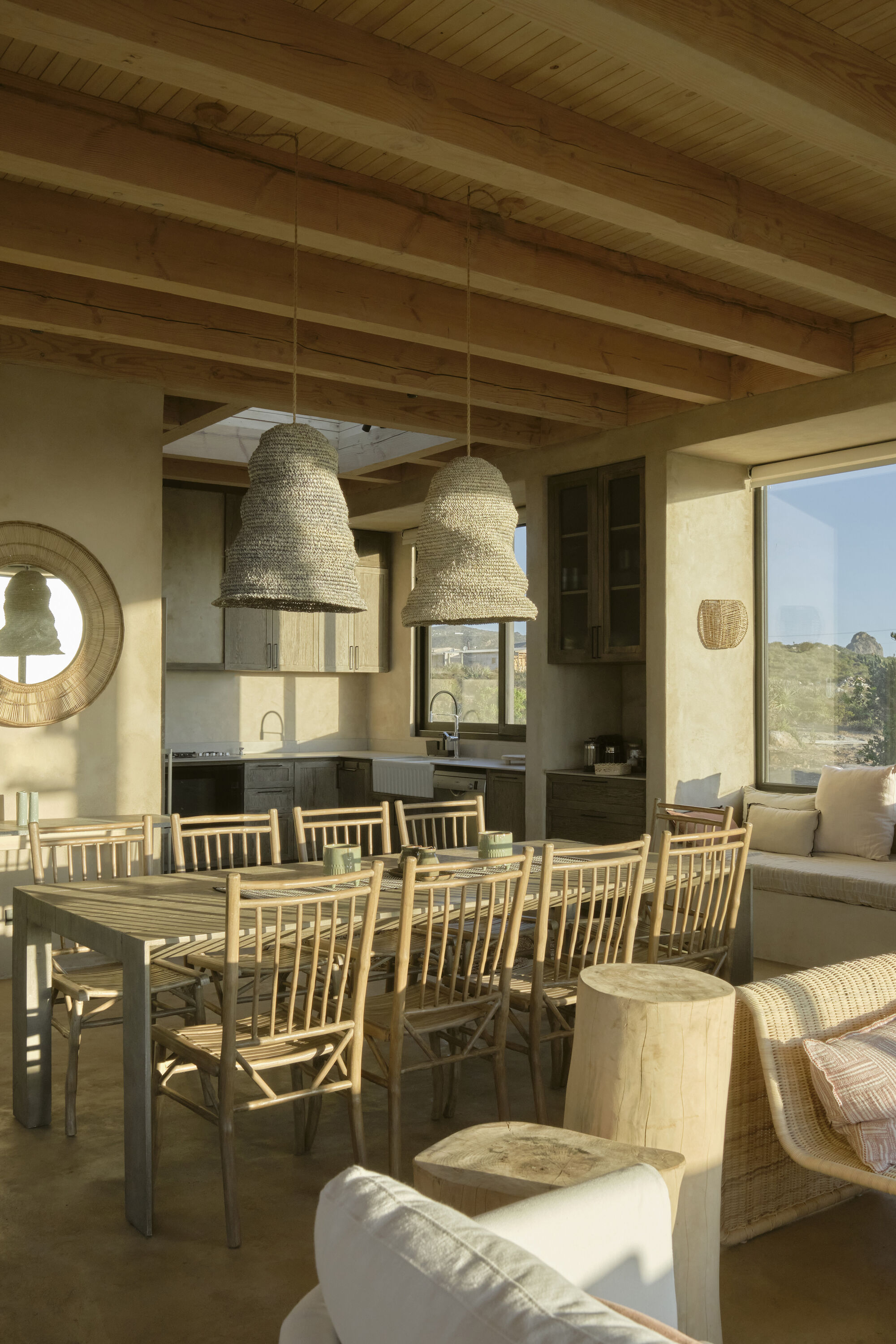 Dining area with a wooden table, bamboo chairs and woven pendant lamps under exposed beams, lit by low sun from the window
