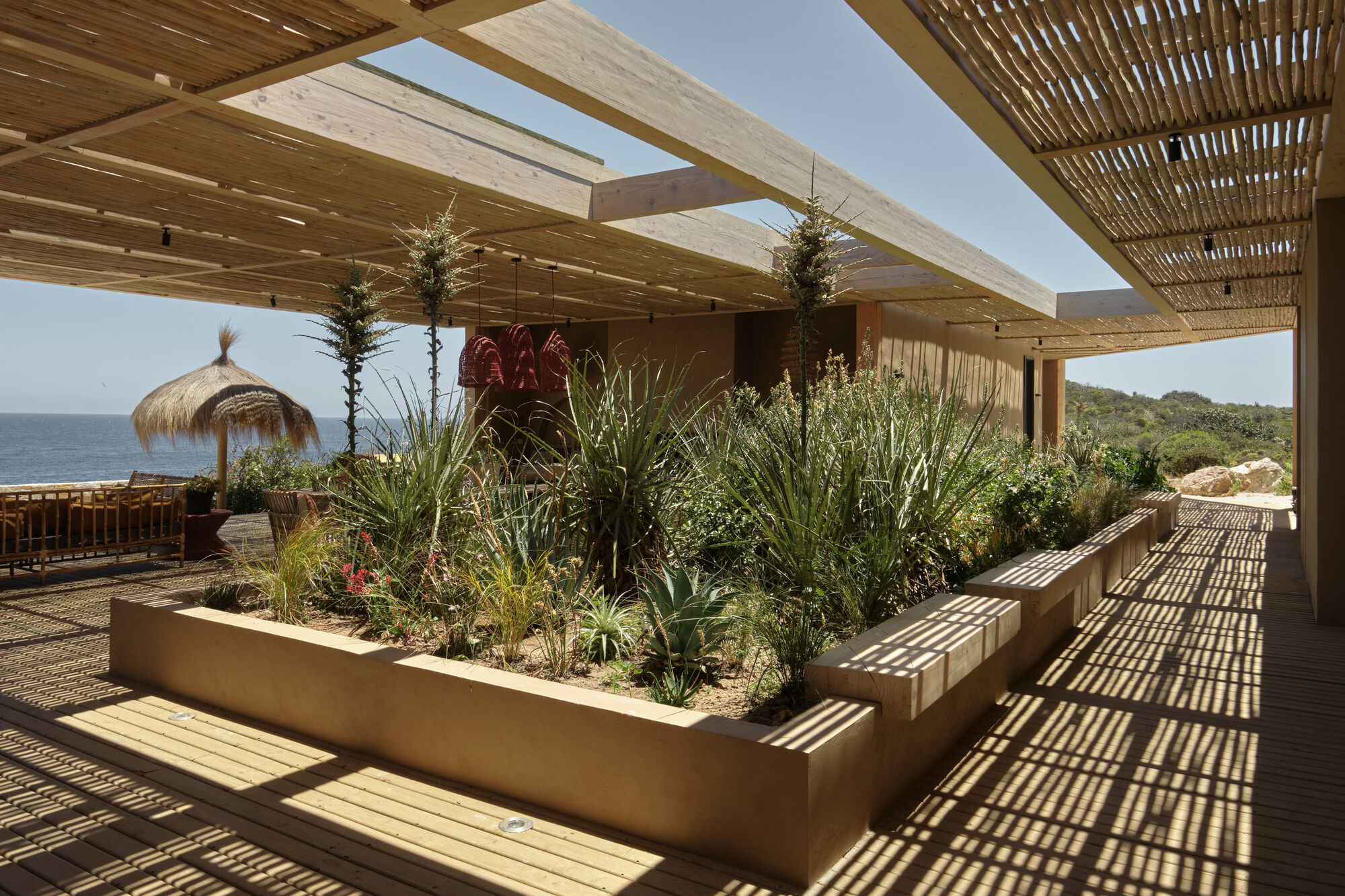 Raised planter with built-in benches and tall grasses beside a reed-screened pergola, casting striped shadows on the deck toward the ocean