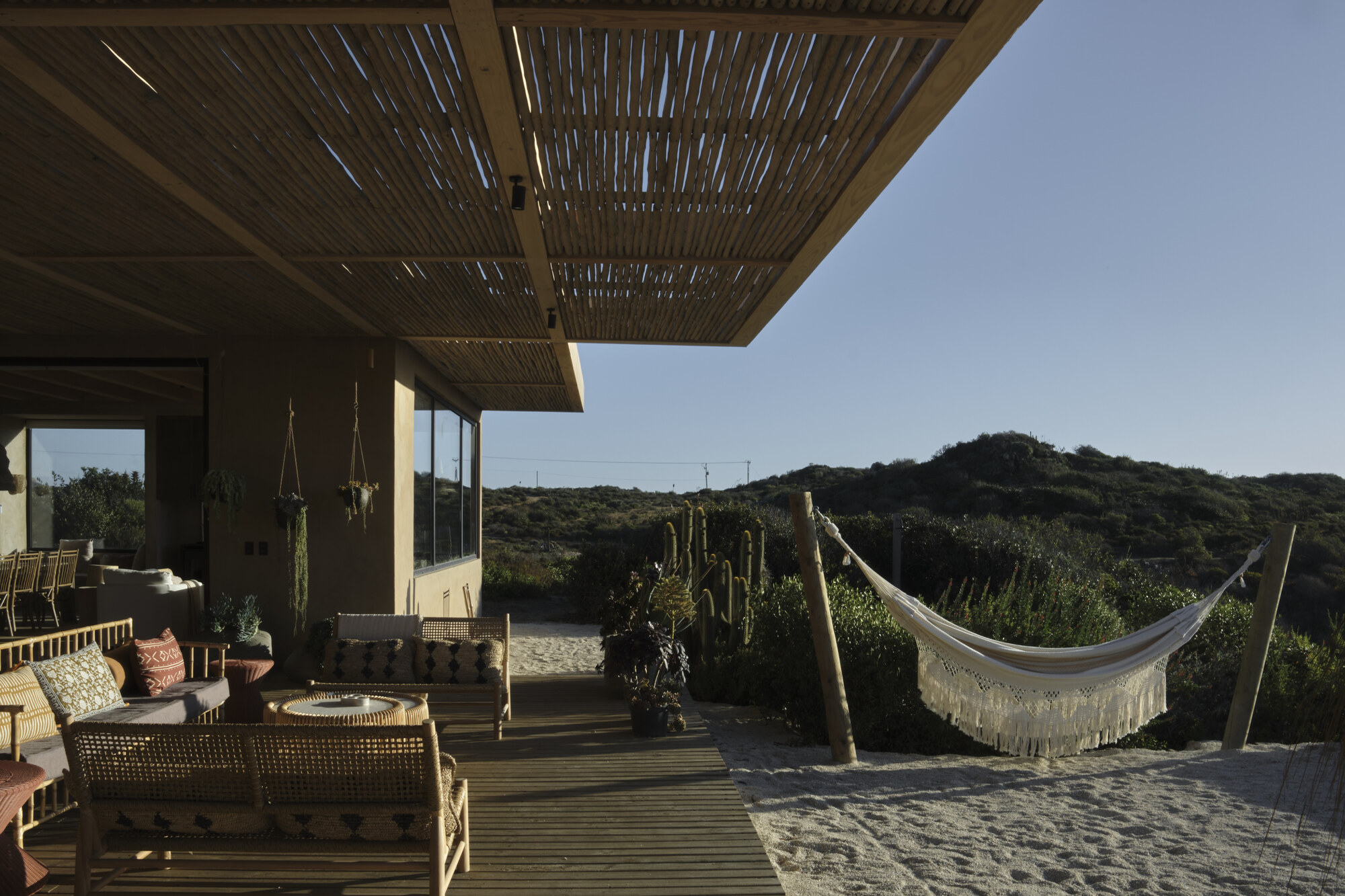Veranda with wicker seating under a slatted roof, opening onto a sandy yard with a fringed hammock and cacti in clear evening light