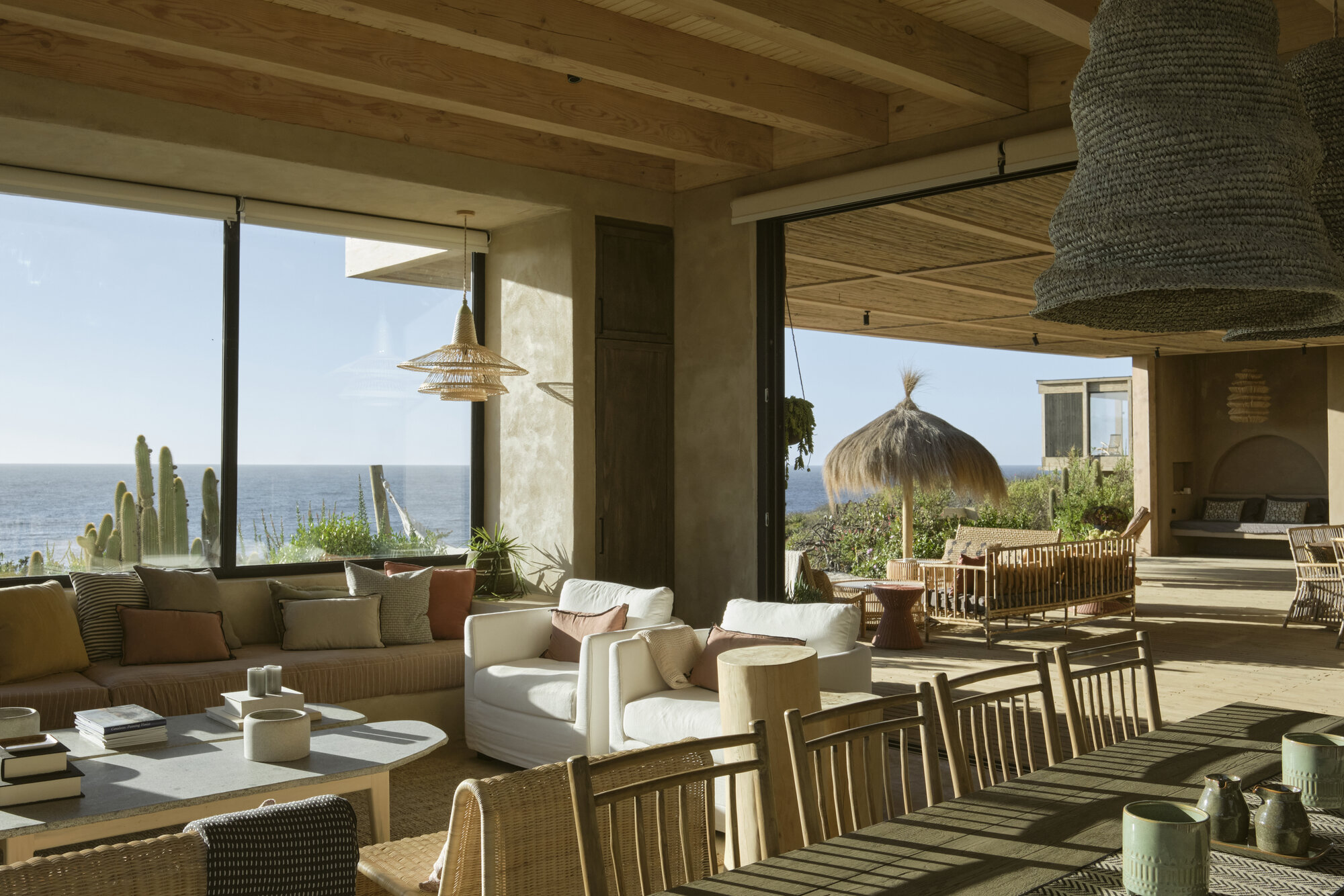 Open lounge and dining area with woven pendant lamps, white armchairs and a thatched umbrella facing the sea under clear daylight
