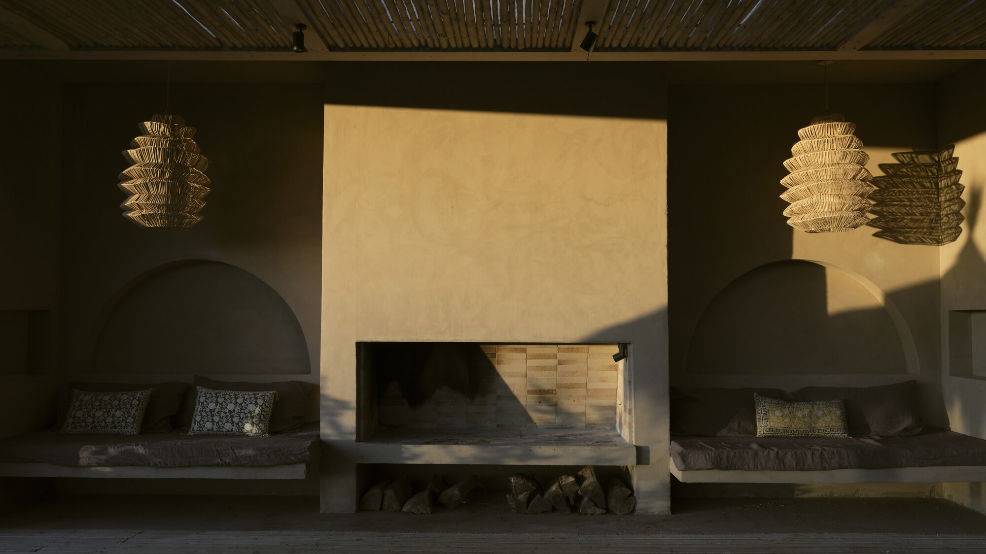 Plastered fireplace wall flanked by arched built-in benches and woven pendant lamps, lit by low golden evening sun