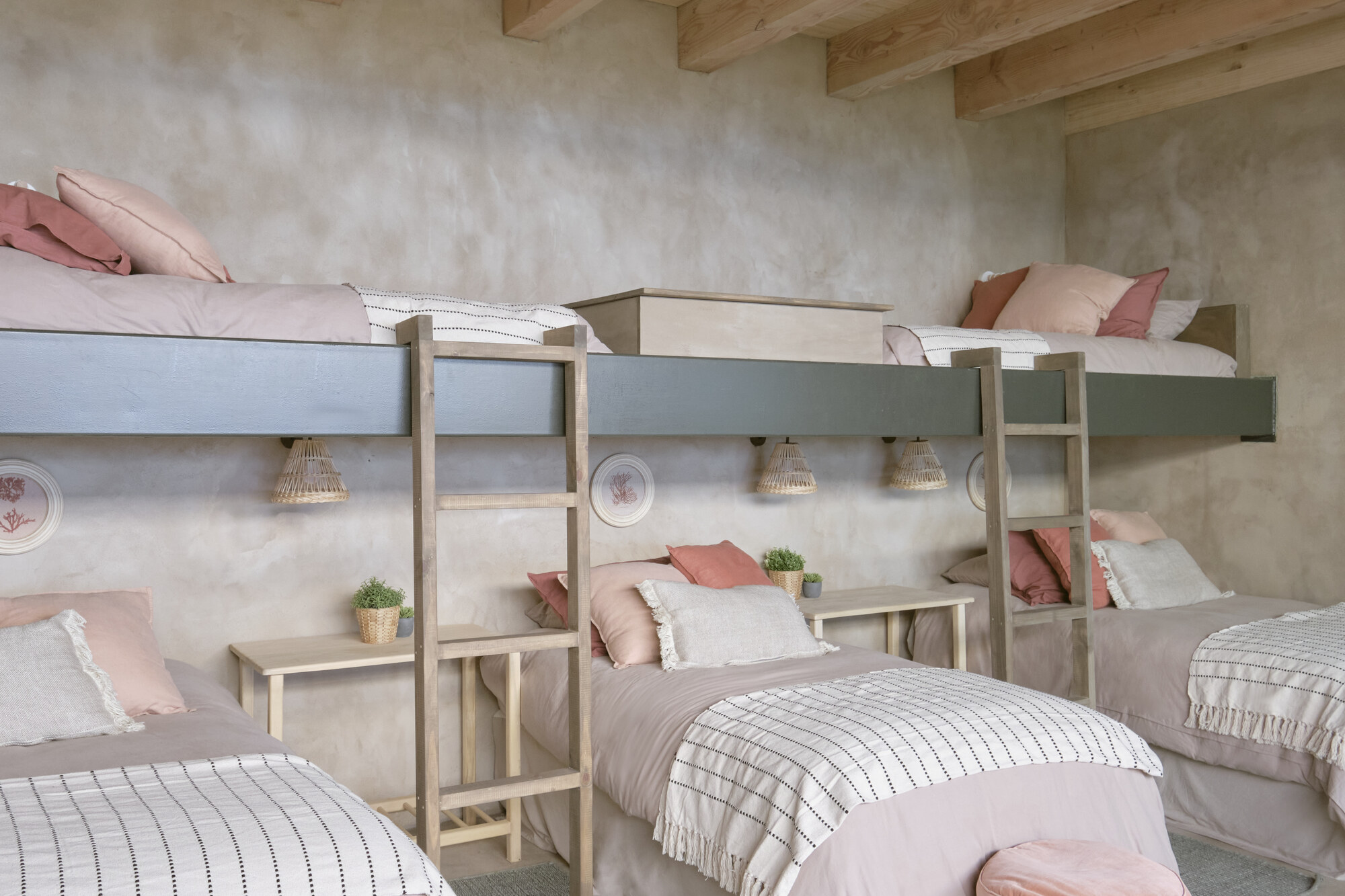 Bunk room with grey-fronted upper berths, timber ladders and pink bedding against a rough plaster wall beneath exposed beams