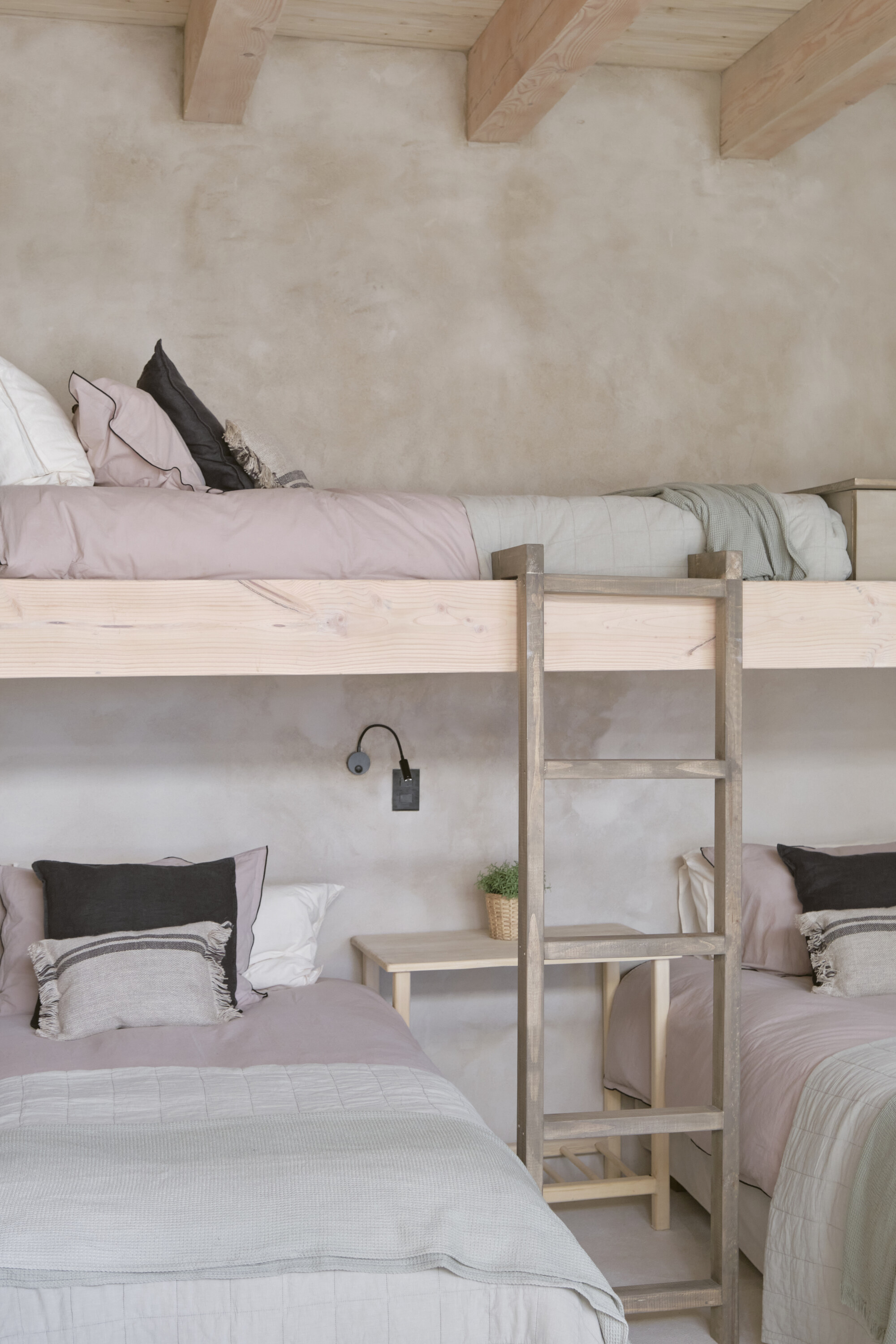 Pale timber bunk bed with ladder and small side shelf against a mottled plaster wall, soft daylight on grey linens