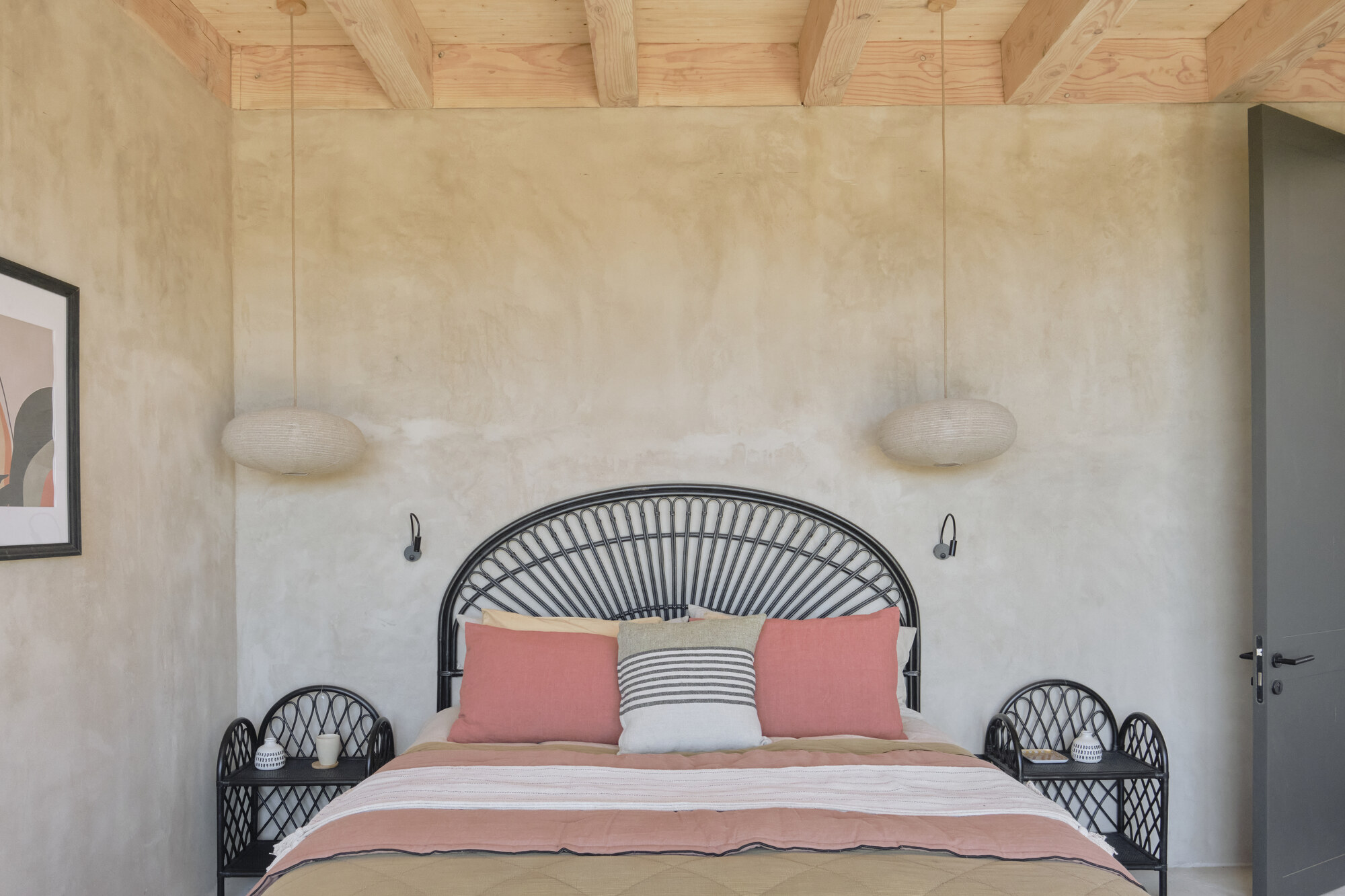 Bedroom with black rattan headboard, matching bedside tables and two round pendant lamps against a plaster wall under timber beams