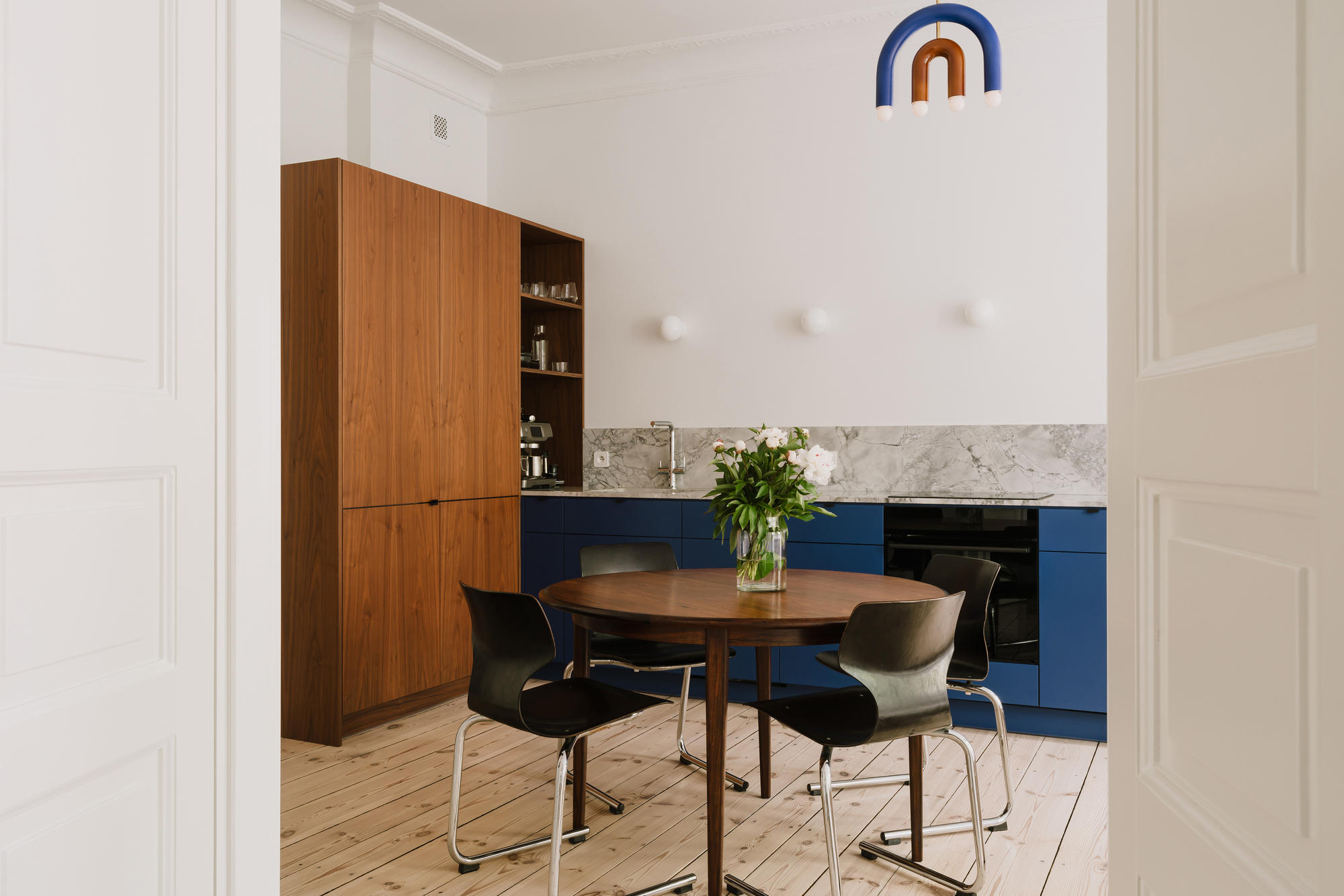 Kitchen seen through a paneled doorway, with a walnut cabinet, blue base units and a round dining table in daylight
