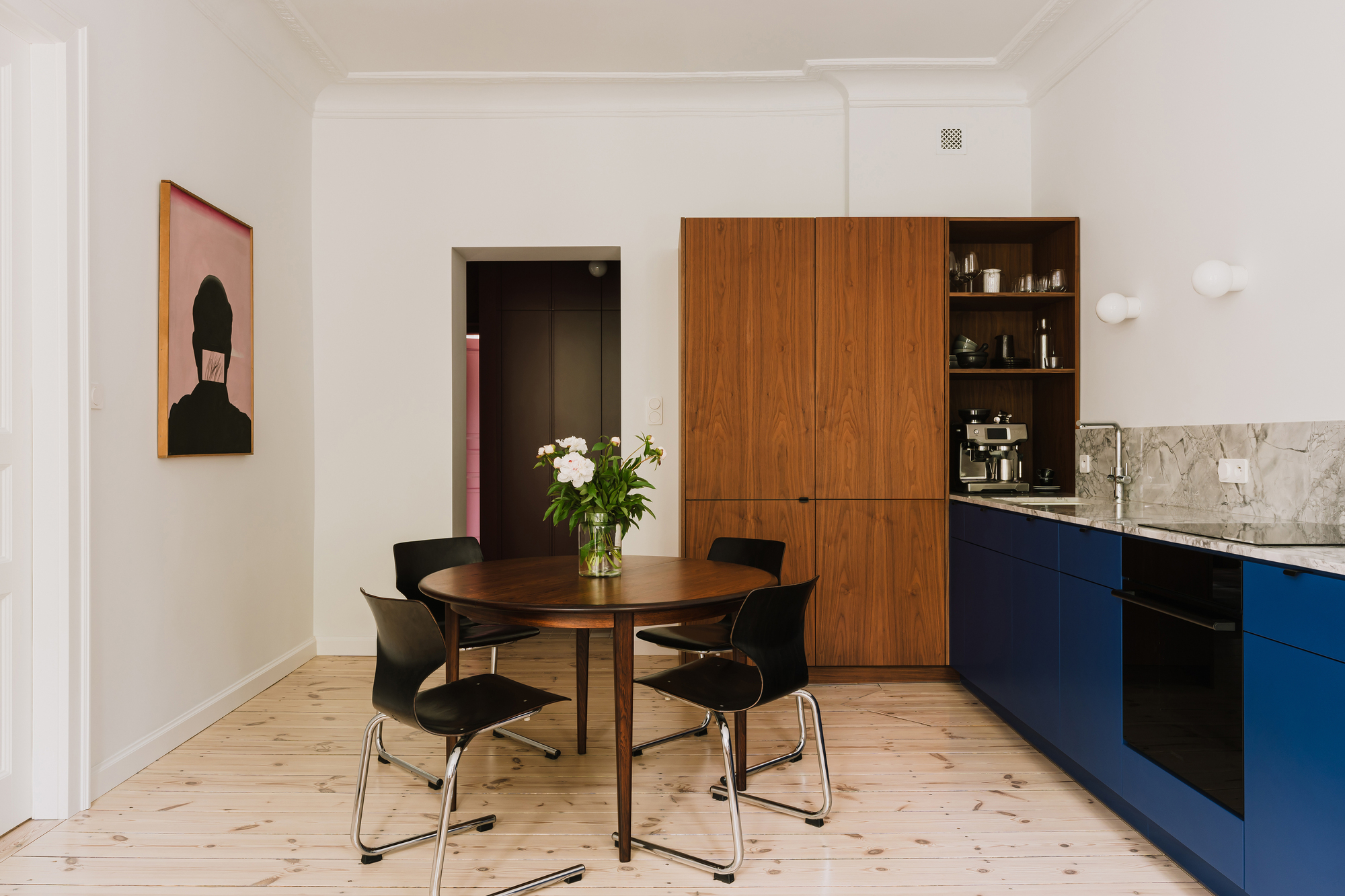 Round walnut table and black chairs beside a blue kitchen run and tall walnut cabinet, with a framed portrait on the wall