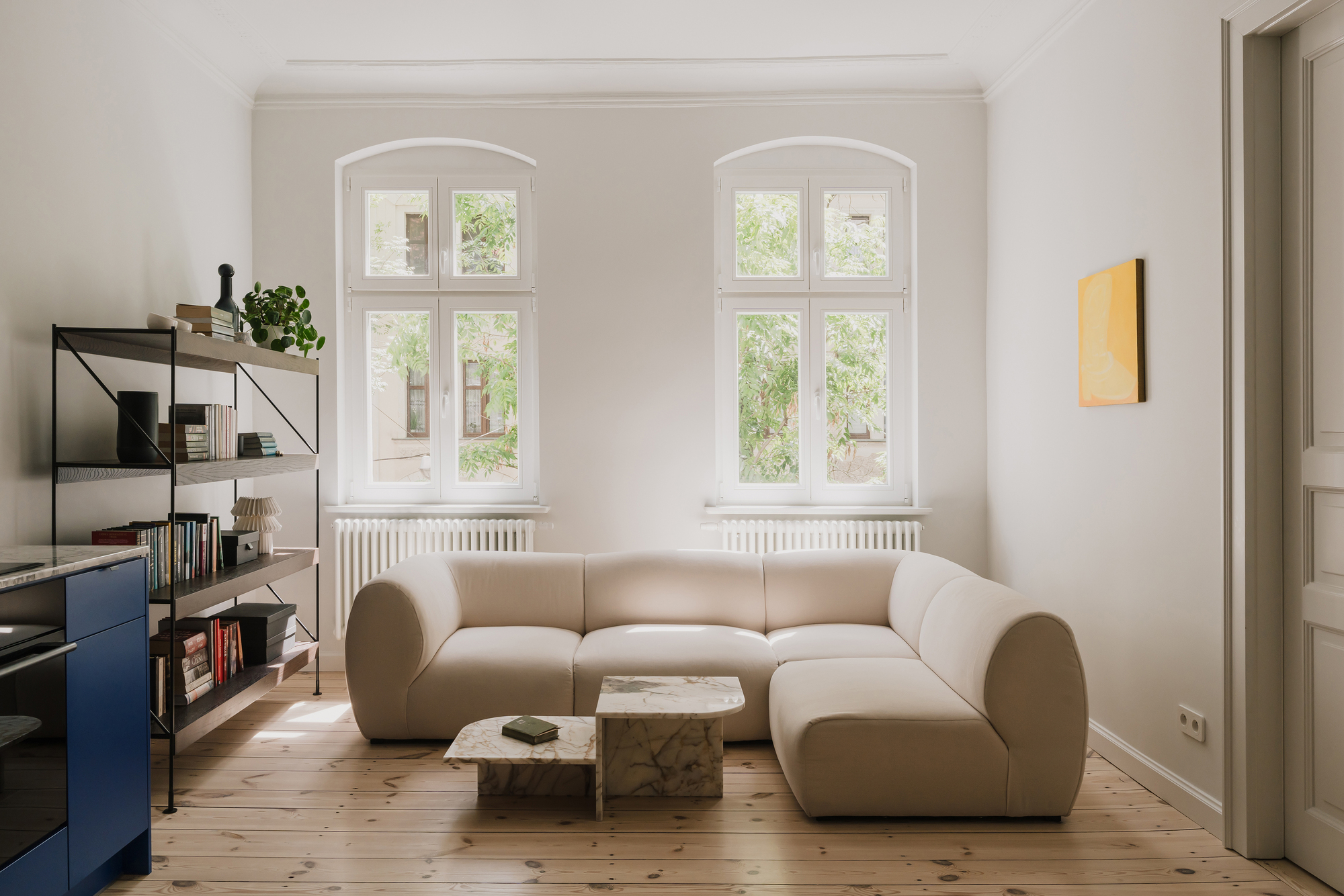 Cream curved sectional sofa and marble coffee tables below two arched windows showing green summer foliage