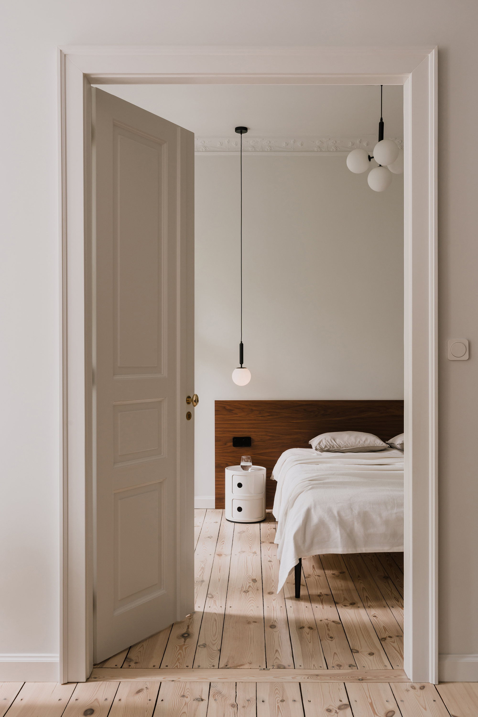 Open paneled door framing a bedroom with a walnut headboard, white round side table and pendant globe lights in soft daylight