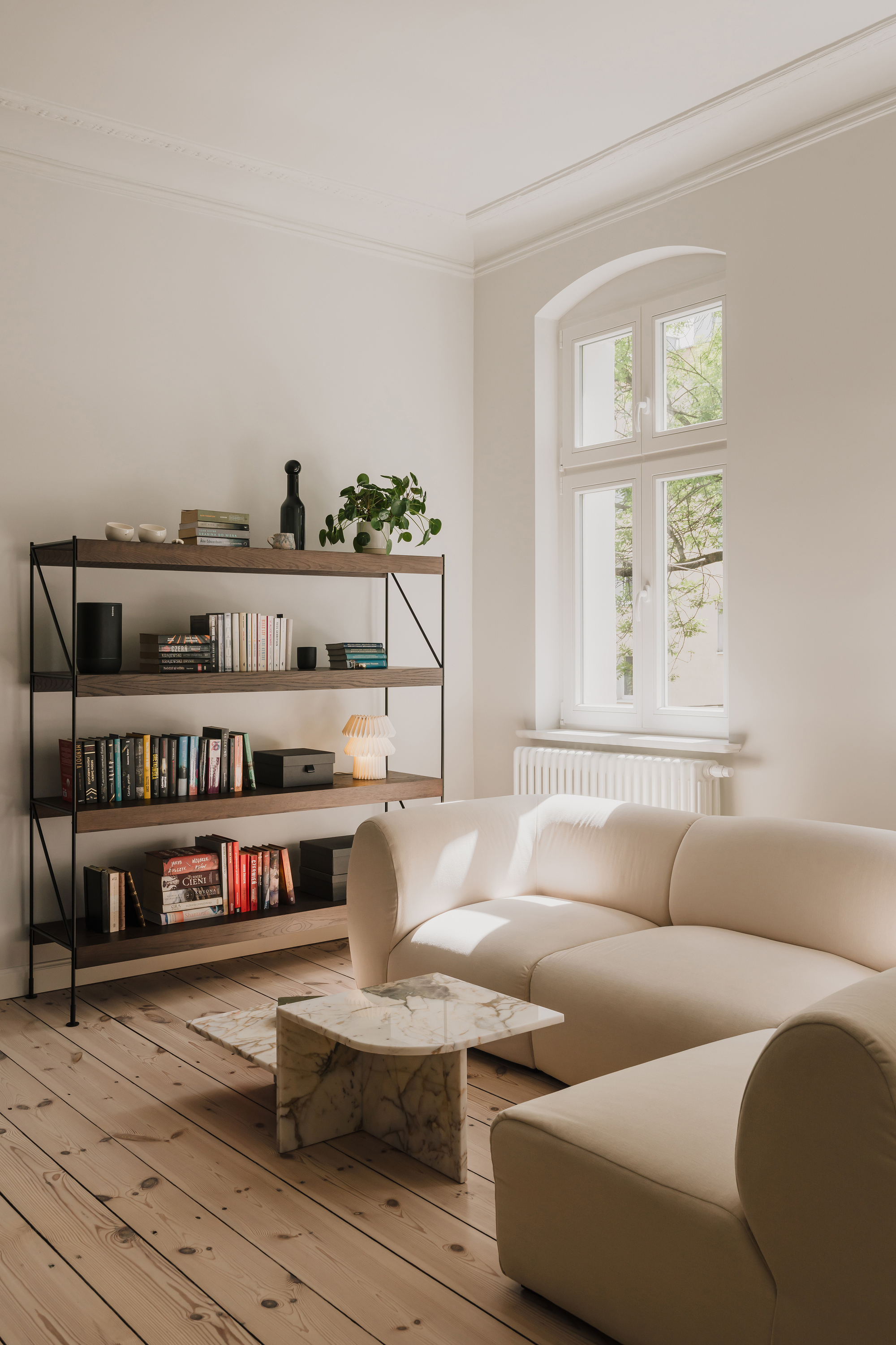 Open timber shelving with books and a potted plant beside a cream modular sofa and marble side table in afternoon sun