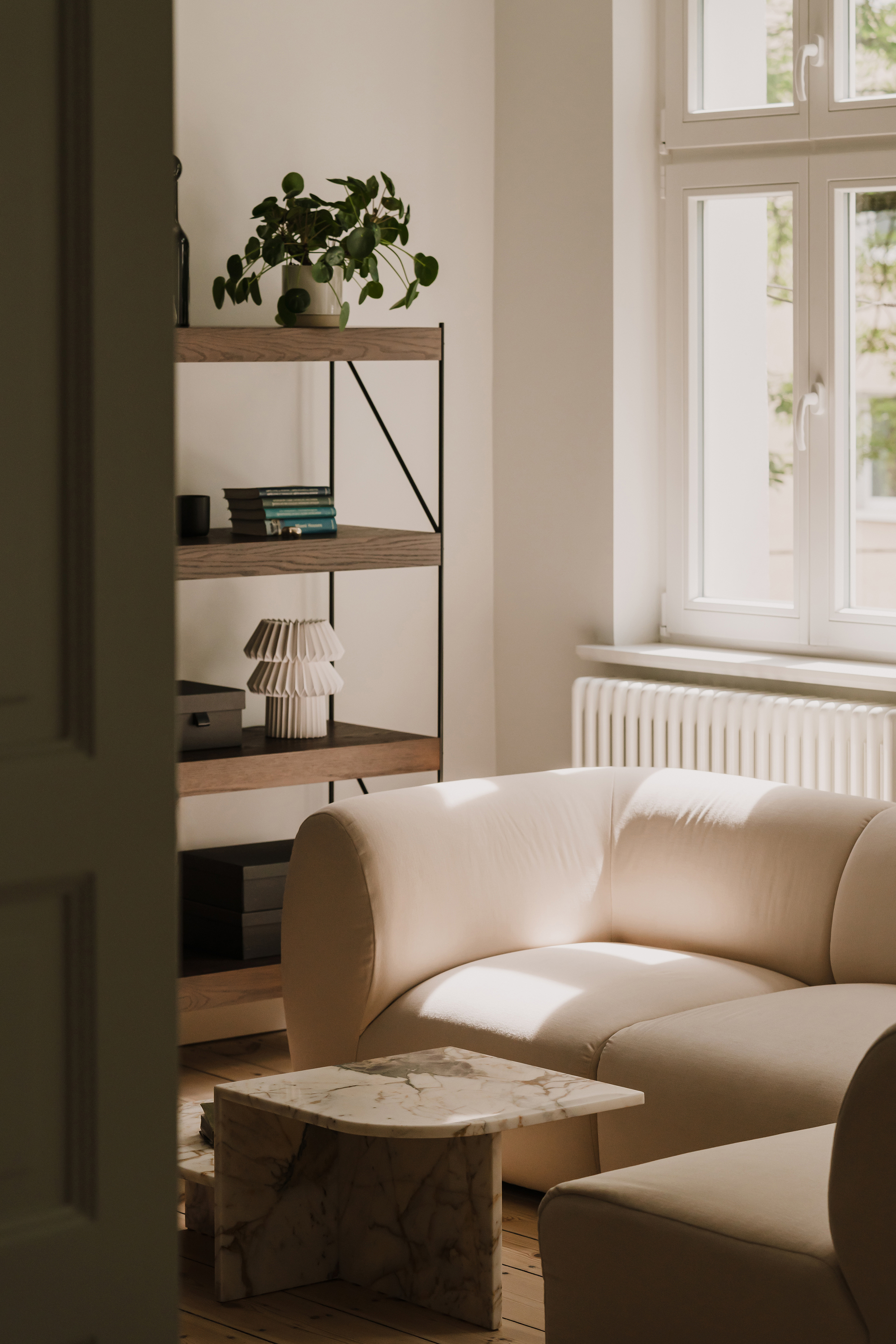 View past a door toward a shelf, pleated lamp and curved cream sofa lit by low sunlight from a tall window