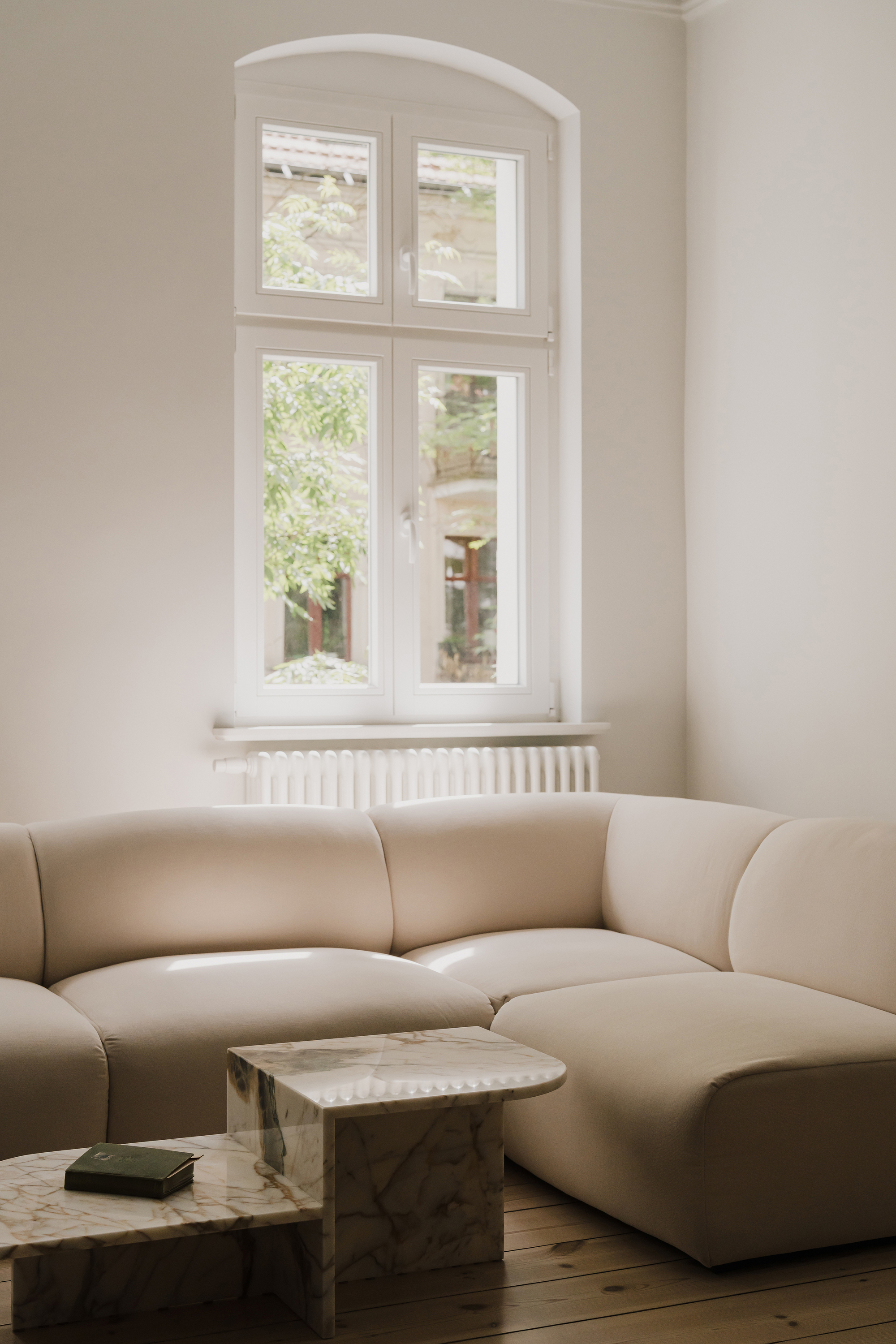 Curved cream sofa and stacked marble tables beneath an arched white window framing green leaves outside