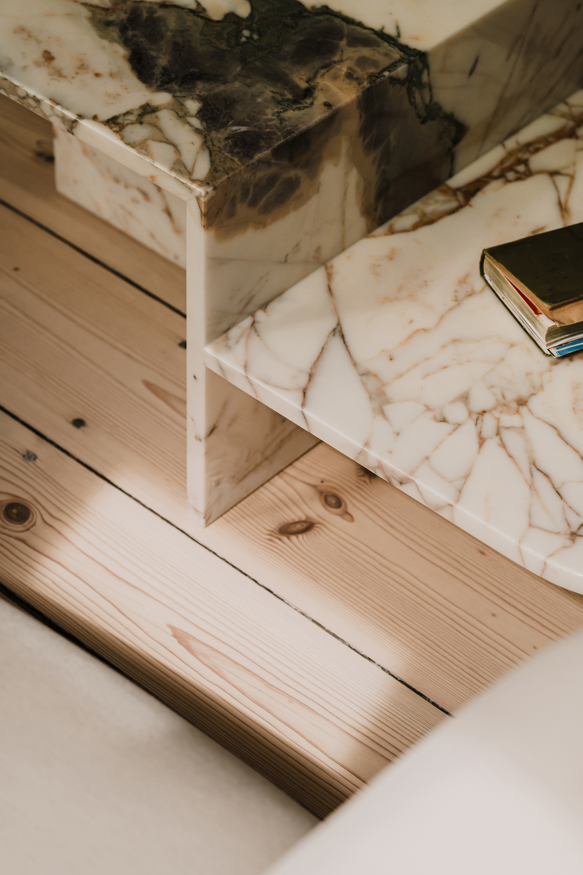 Close-up of veined marble table edges resting on pine floorboards, with a small book catching a patch of sunlight