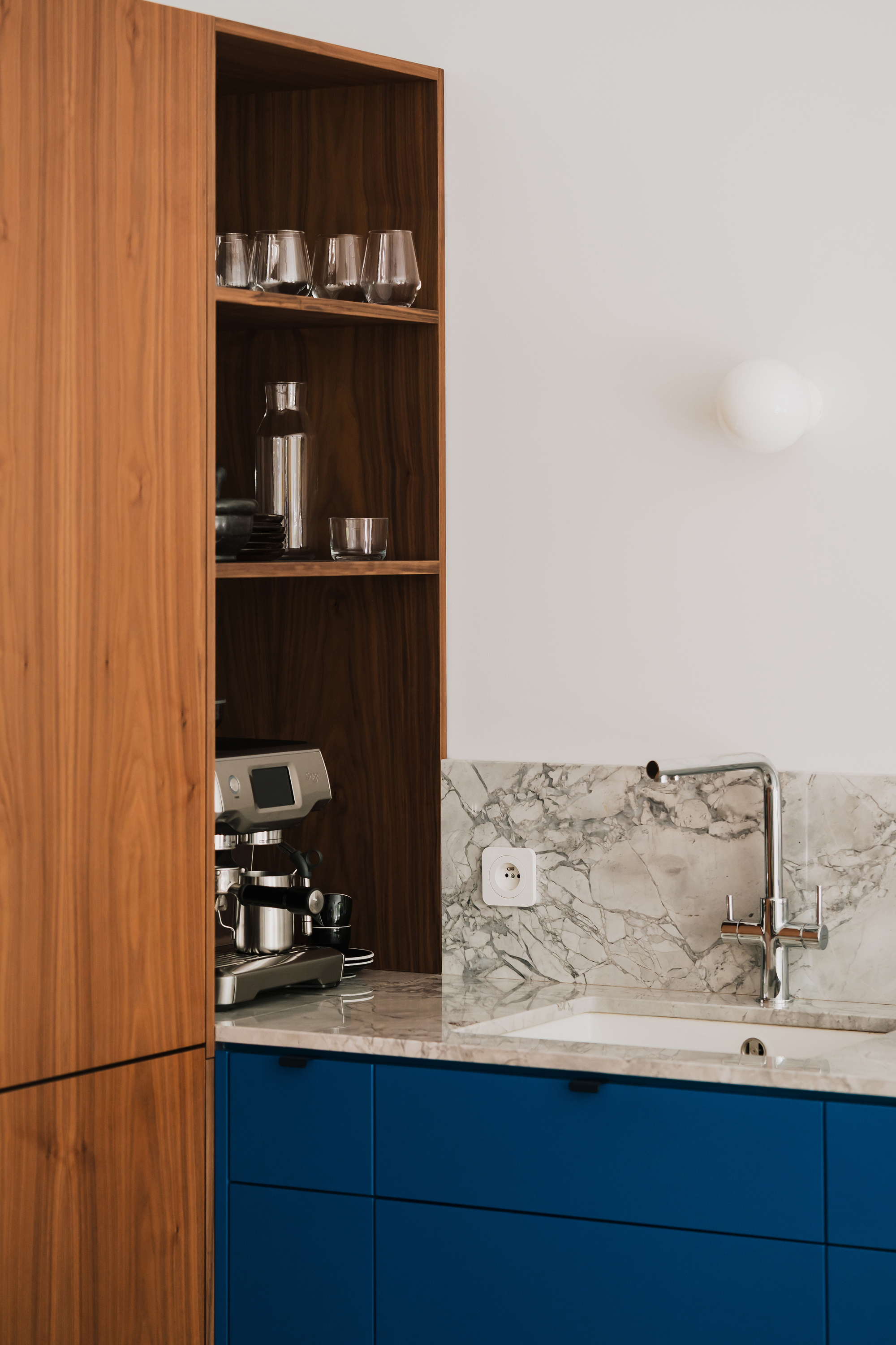 Walnut open shelving with glasses and an espresso machine above blue cabinet fronts and a marble backsplash with sink