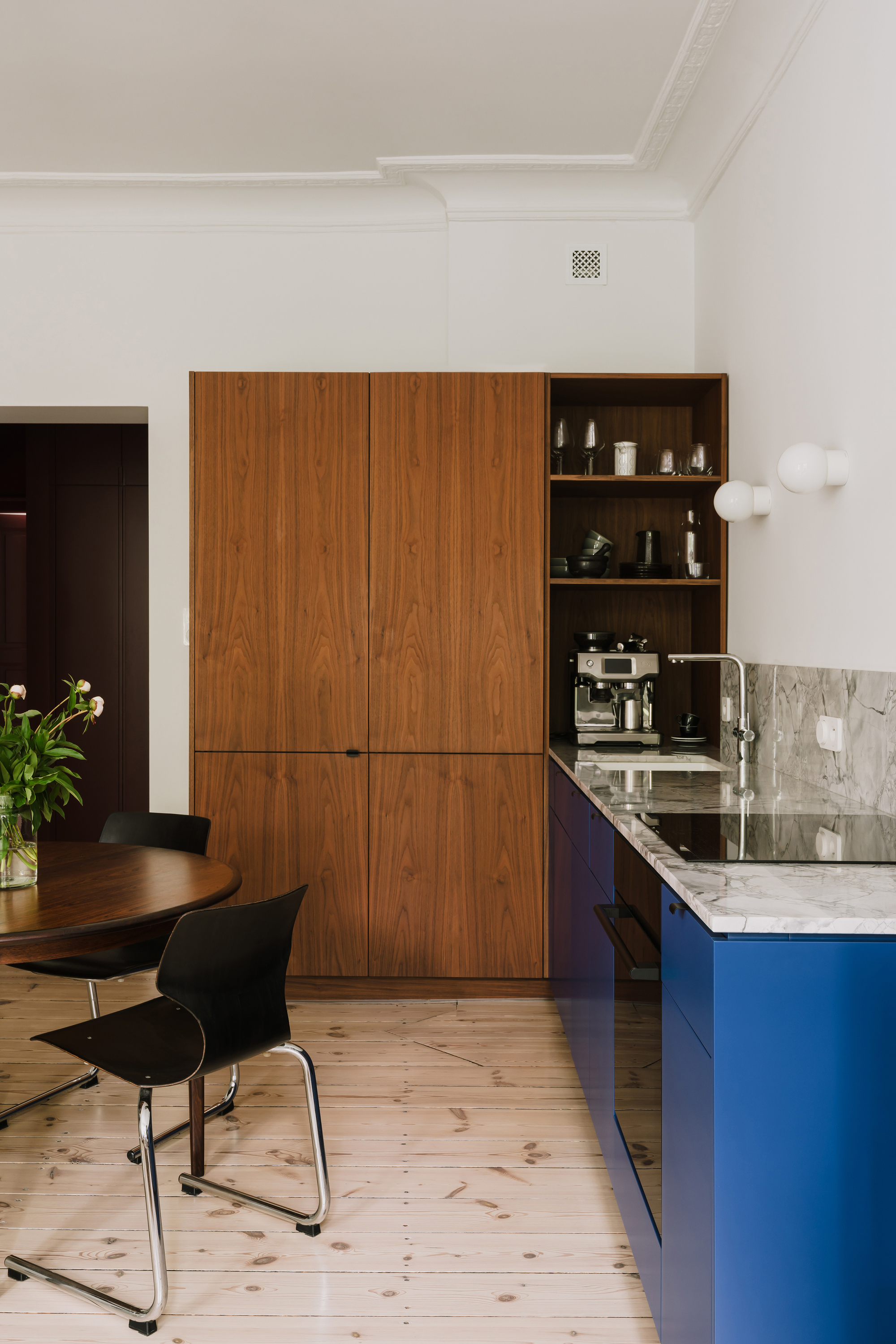 Kitchen corner with walnut tall cabinets, blue base units and black cantilever chairs at a round table beside tulips