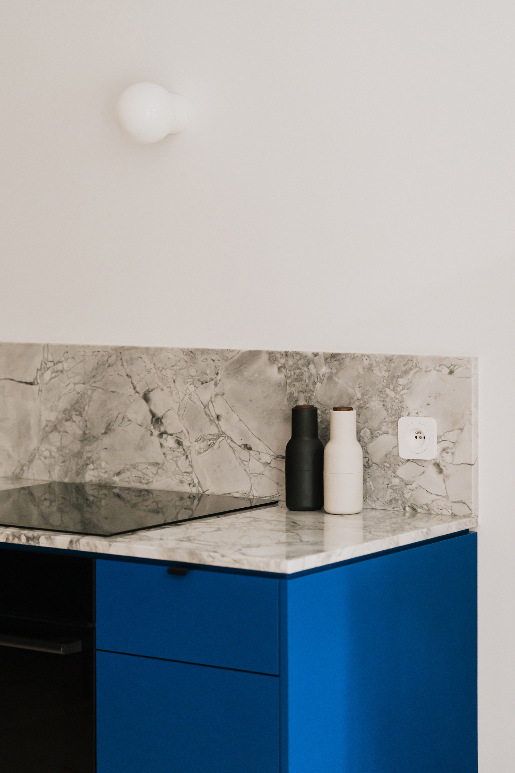 Corner of a kitchen counter with grey veined marble backsplash, induction hob and two grinders above blue cabinet fronts