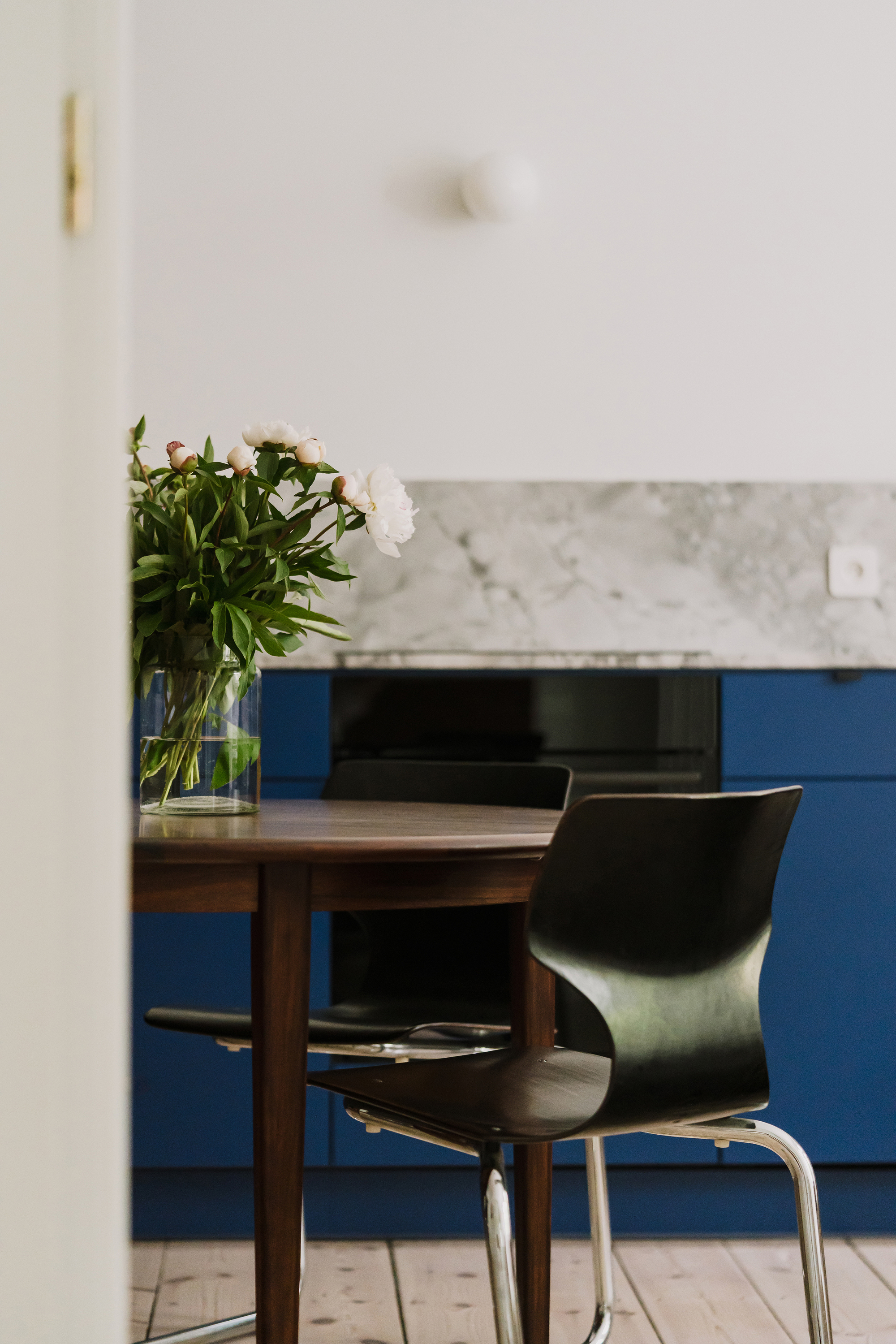 Round walnut dining table with black bentwood chairs and a vase of white peonies in front of blue kitchen cabinets
