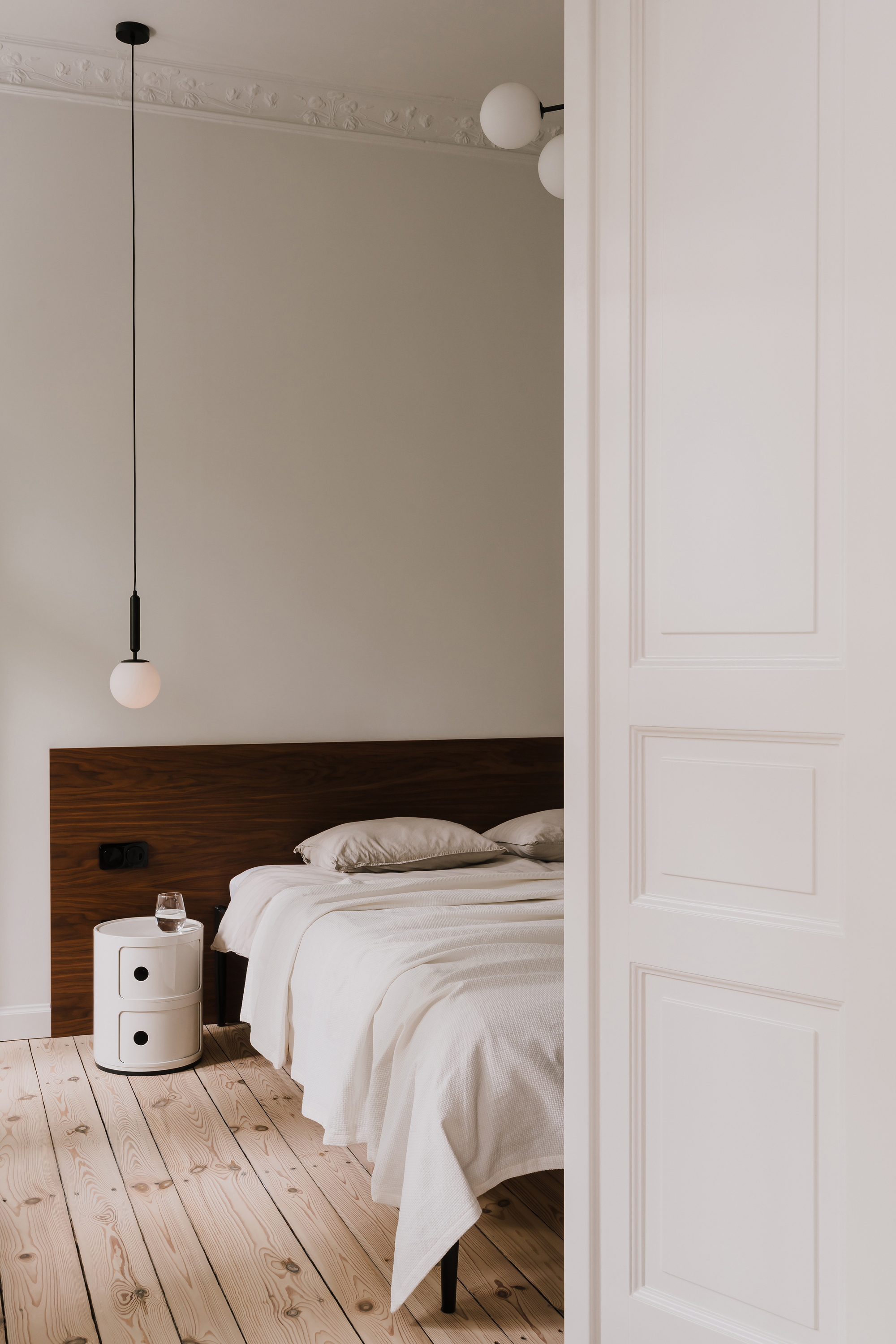 Bedroom seen through a paneled white door, with walnut headboard, round pendant lamp and a white side unit on pine floorboards