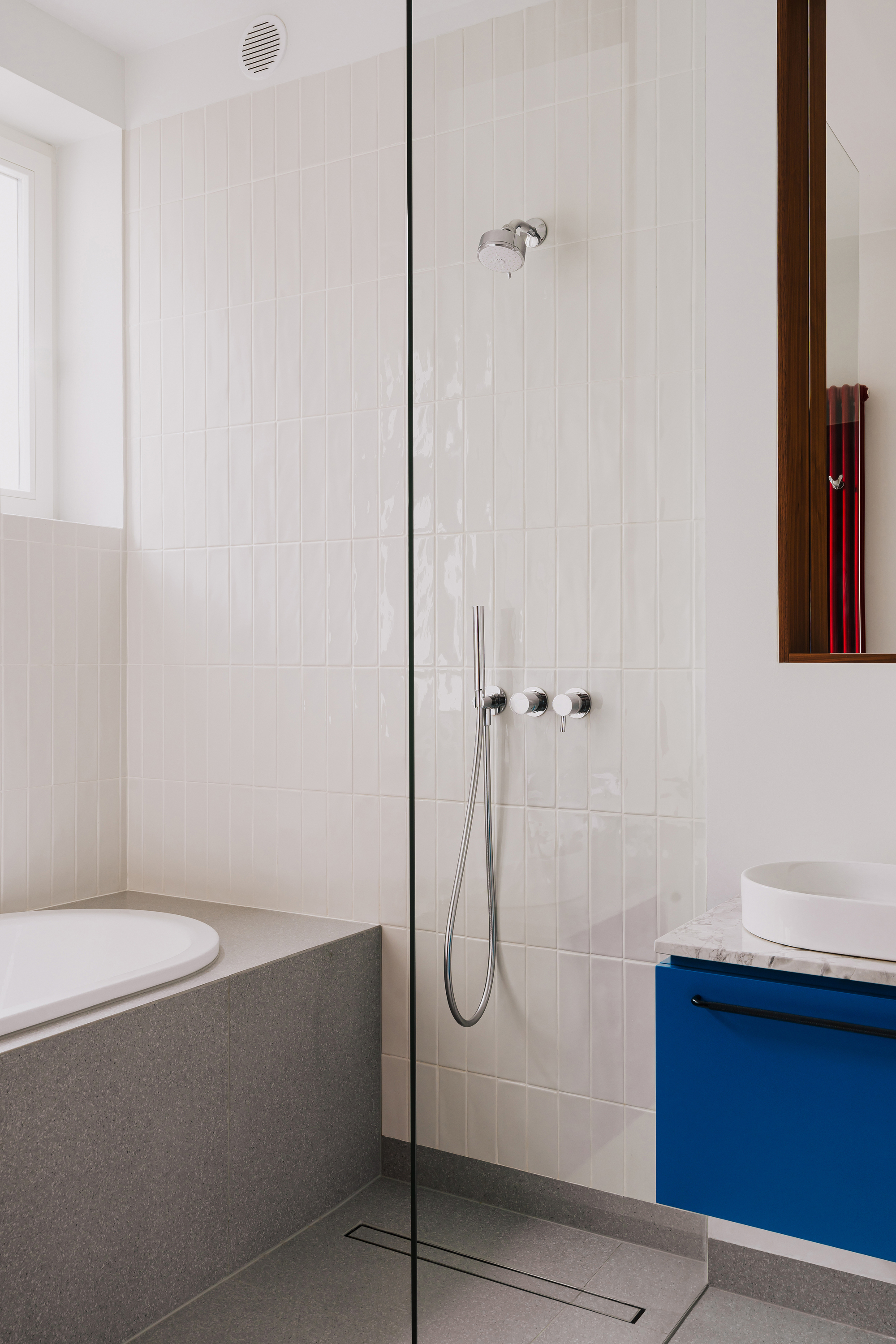 Shower behind a glass panel with vertical glossy white tiles, chrome fittings and a grey-clad bathtub beside a window