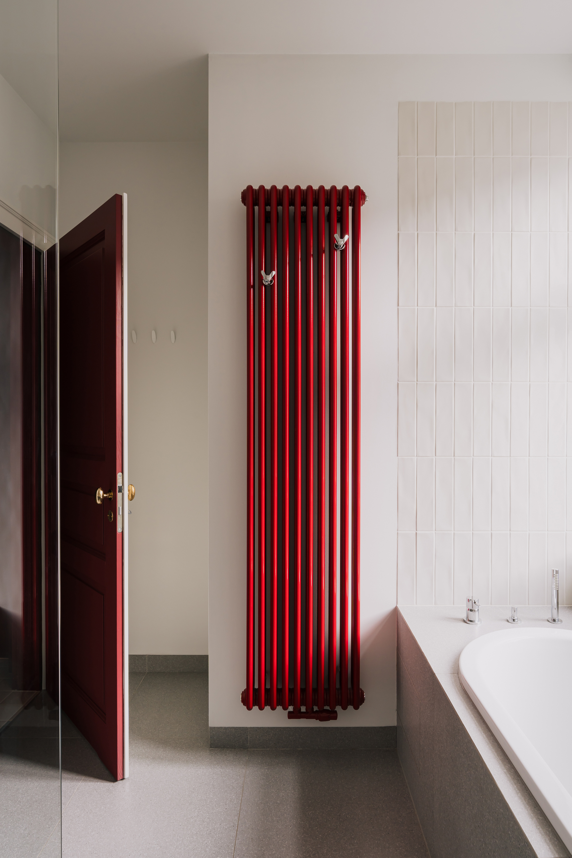 Tall red column radiator with two hooks beside an open maroon door, next to white tiles and a bathtub edge