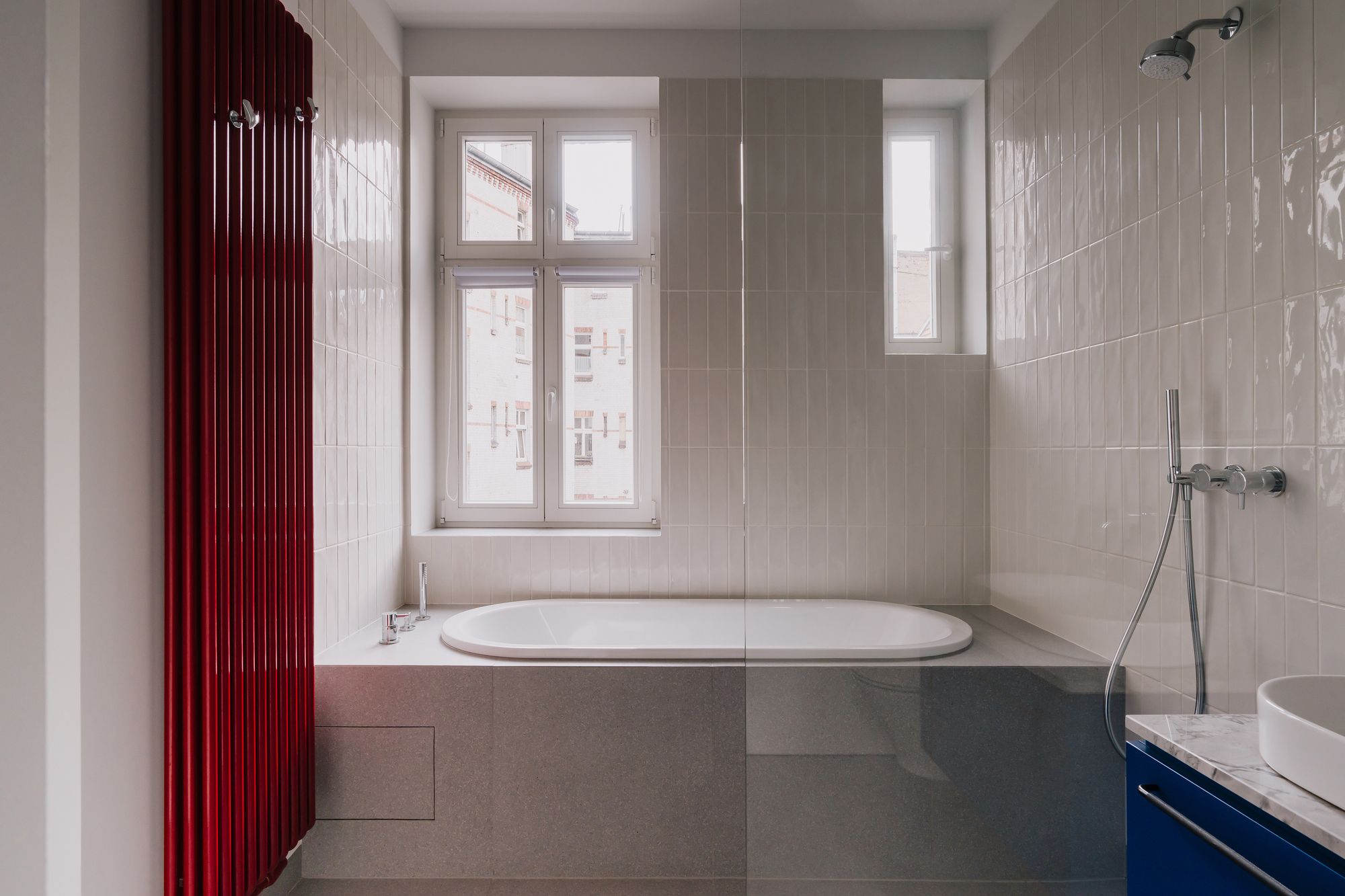 Bathroom with a built-in oval tub below a window, vertical white glazed tiles and a tall red radiator in soft daylight