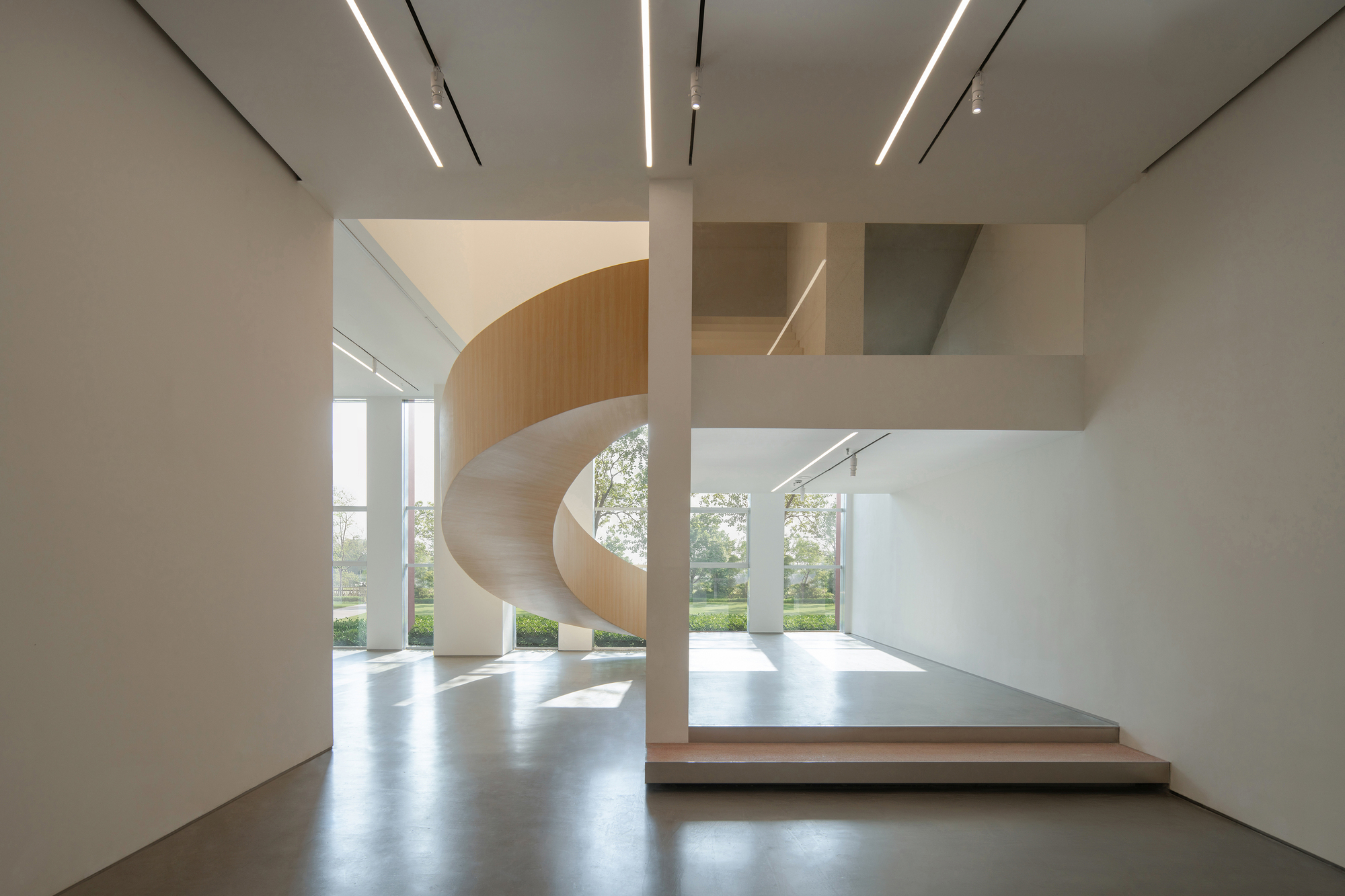 Spiral timber-clad staircase behind a white column in a double-height hall, with sunlight patches on the polished concrete floor